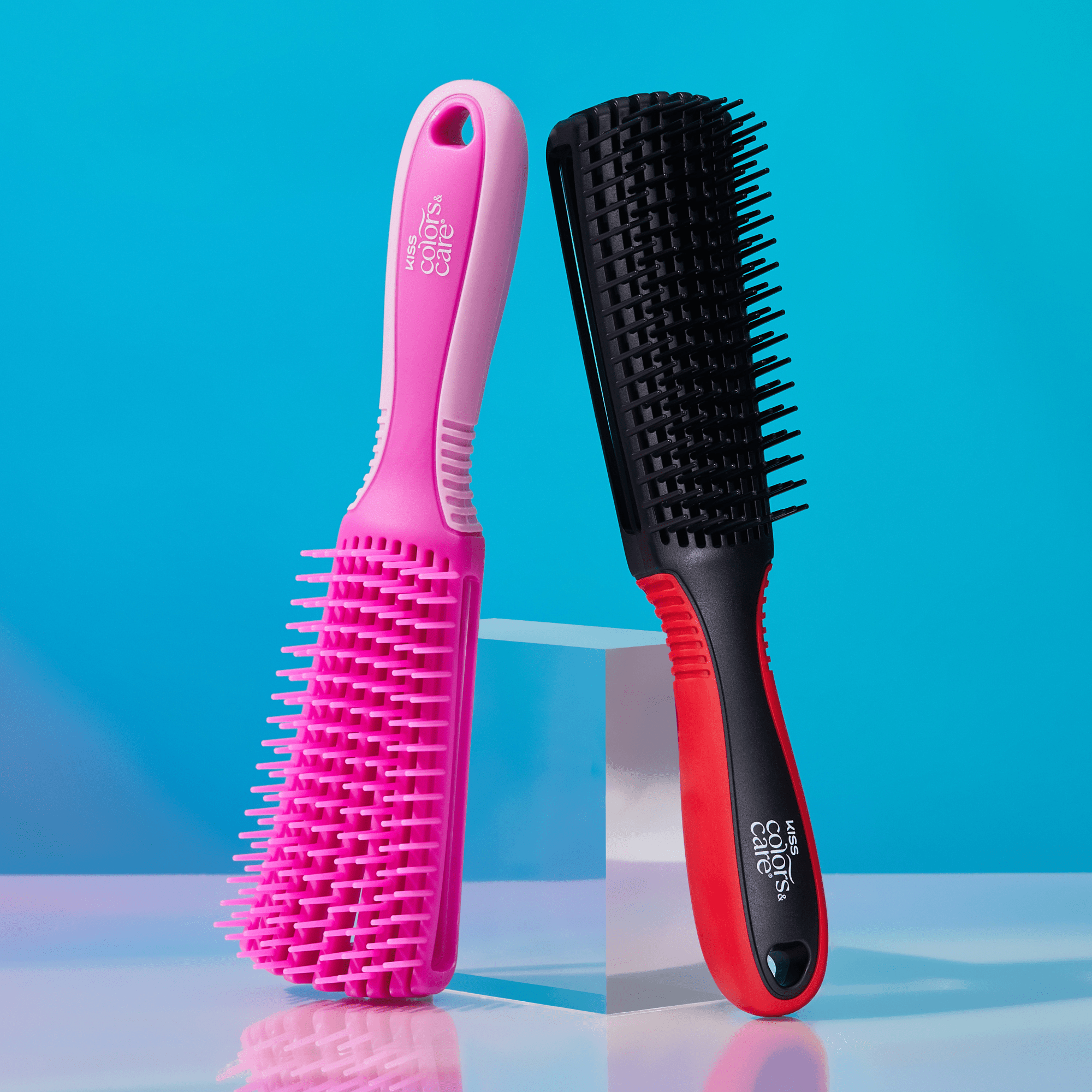 Two KISS Colors &amp; Care Glide &amp; Define Non-Slip Detangling Brushes standing upright on a clear block against a blue background; one brush is pink with light pink grip details, and the other is black with red grip details.