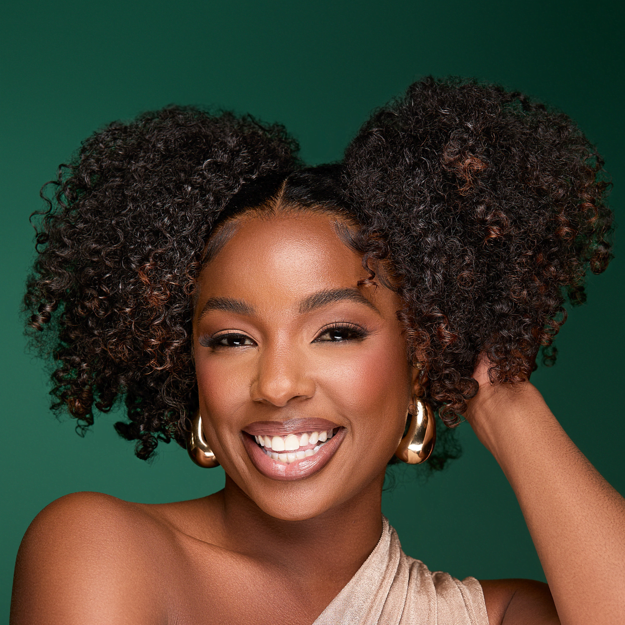 Smiling woman with natural curly hair styled in two puffs, wearing gold hoop earrings and a beige one-shoulder top against a green background.