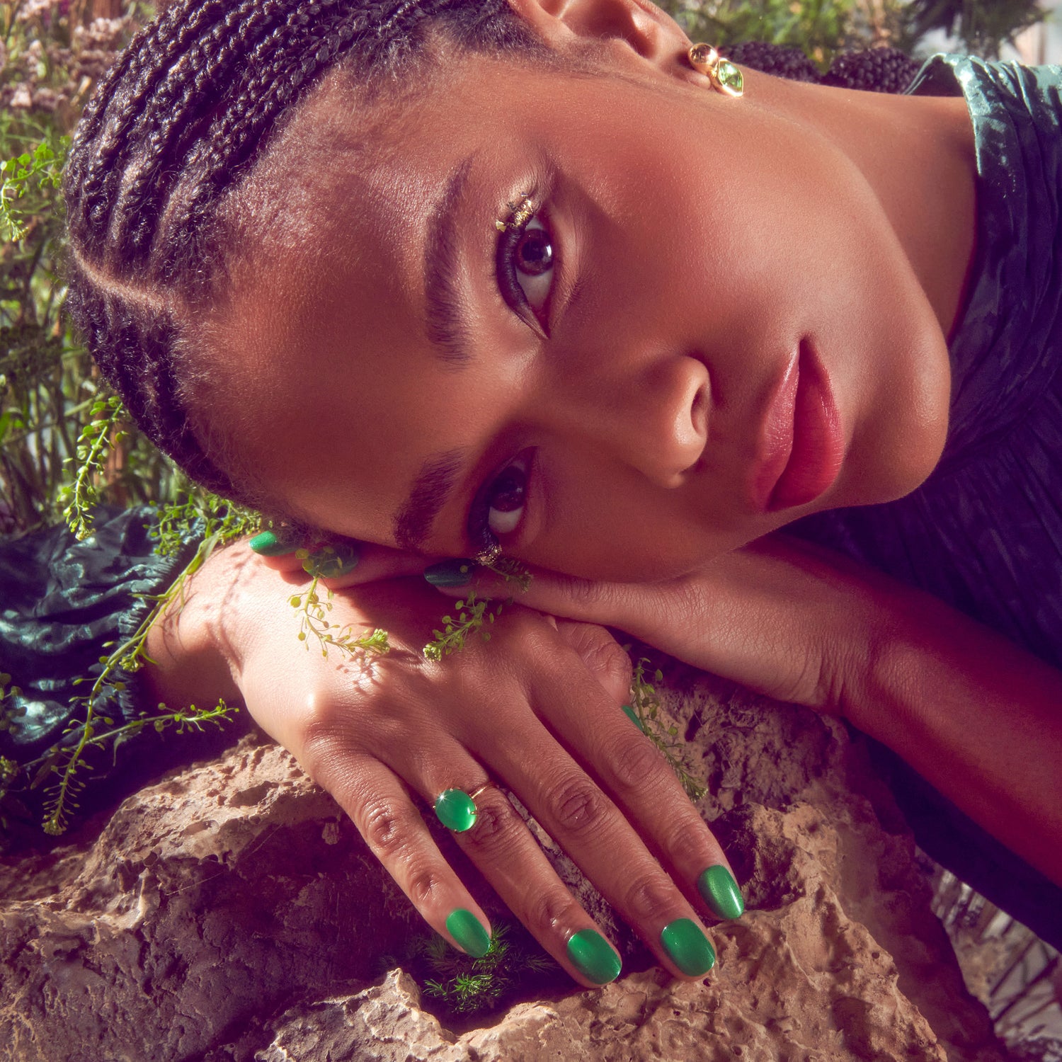 Close-up of a woman with braided hair resting her head on her hands over a textured rock, wearing green nail polish and a green ring, showing natural-looking false eyelashes with gold accents.