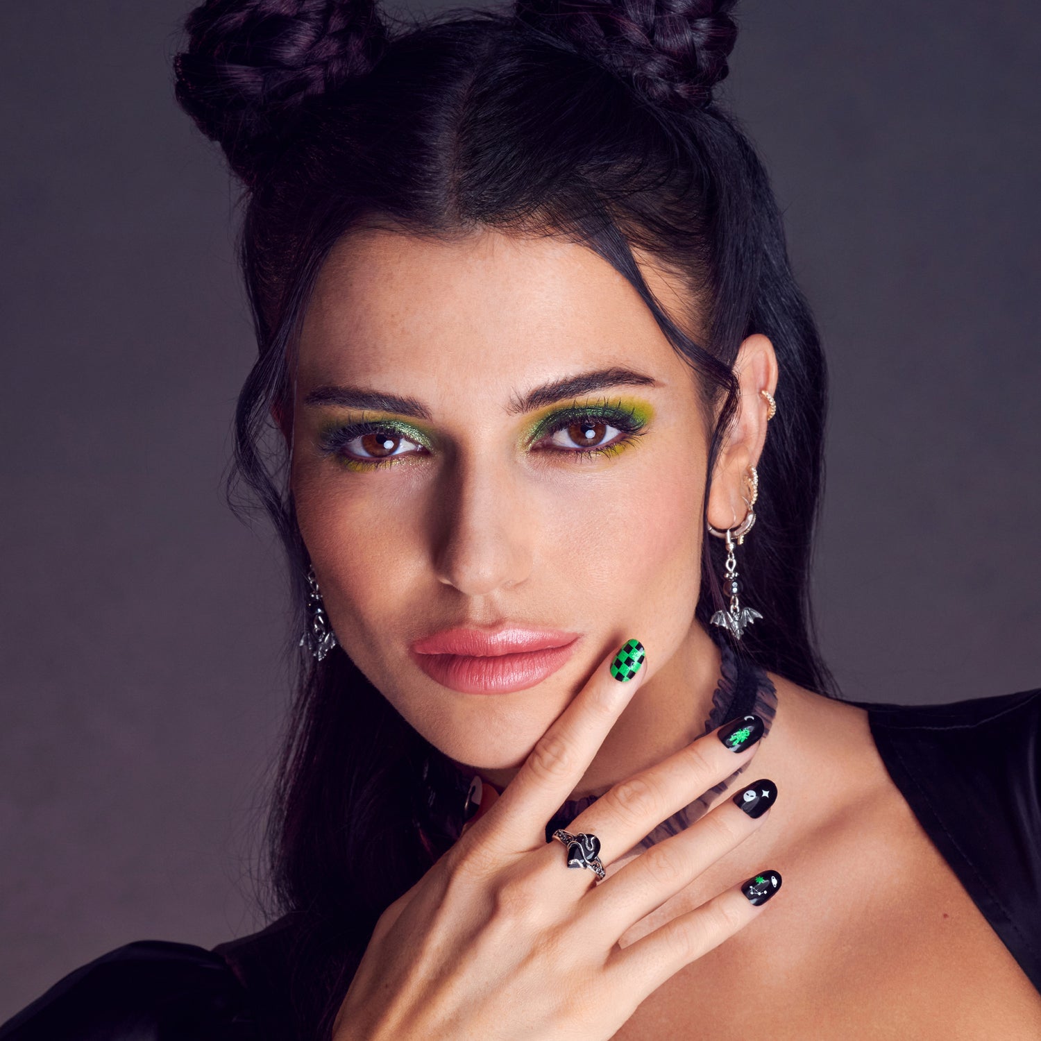 Close-up of a woman with dark hair styled in double buns, wearing light volume false eyelashes, vibrant green eyeshadow, black and green Halloween-themed nail art, and black bat-shaped earrings.
