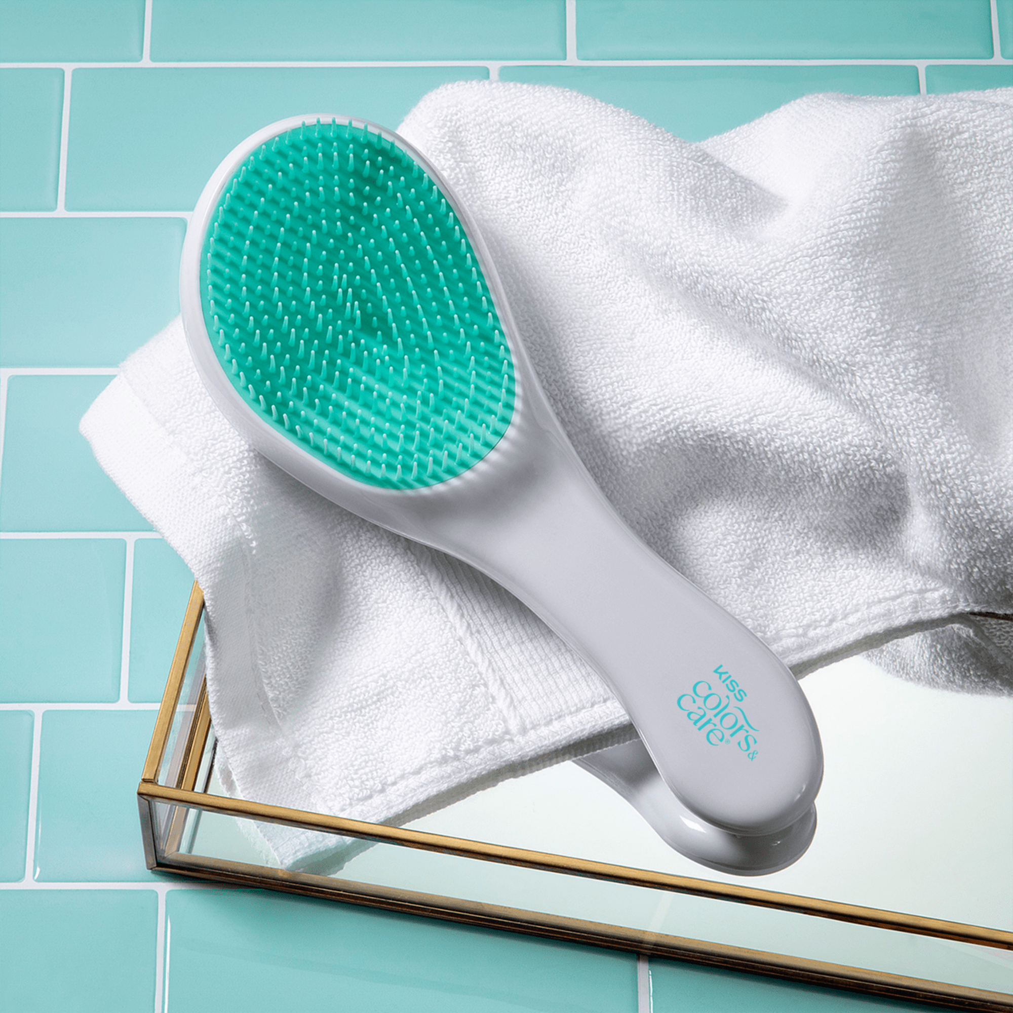 White and teal detangler hairbrush with flexible bristles, lying on a white towel on a mirrored tray over turquoise tiled background, labeled \