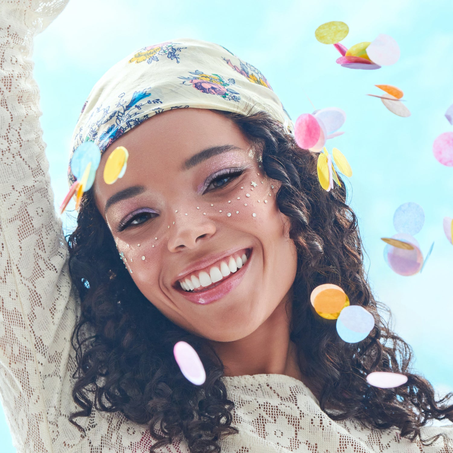 Woman with glitter face art, floral headwrap, colorful ear accessories, smiling against light blue background.