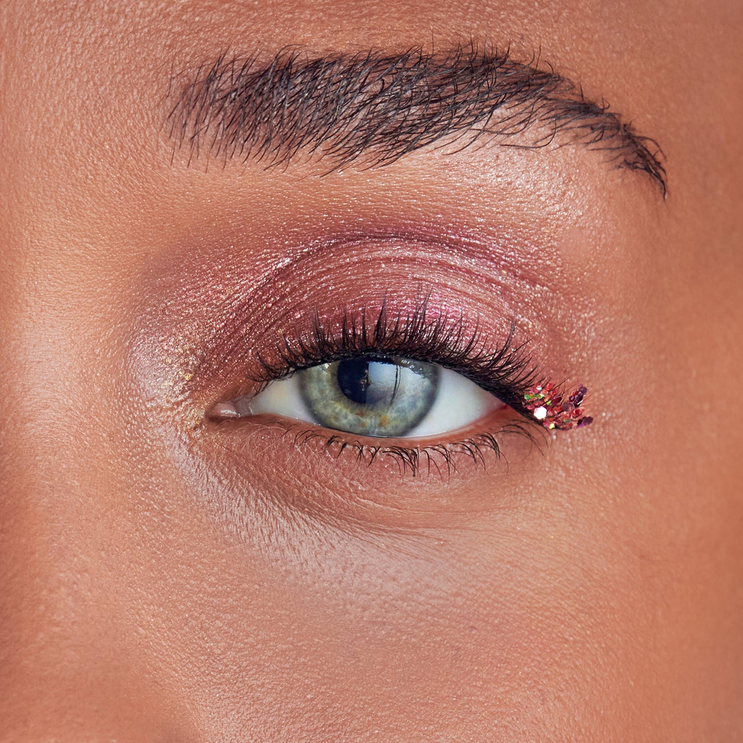 Close-up of an eye with light green iris wearing glittery purple-pink eyeshadow and a small cluster of multicolored glitter pieces on the outer corner of the upper eyelid.