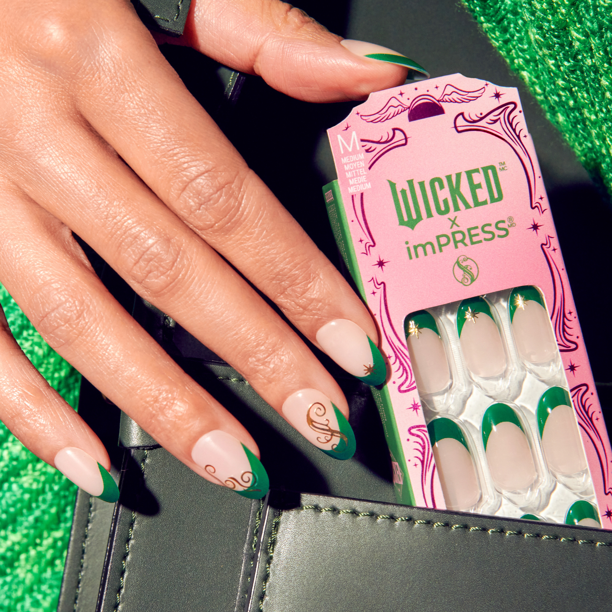 Hand with medium-length nails painted in green French tips and gold swirl accents, holding pink WICKED X imPRESS nail kit labeled \