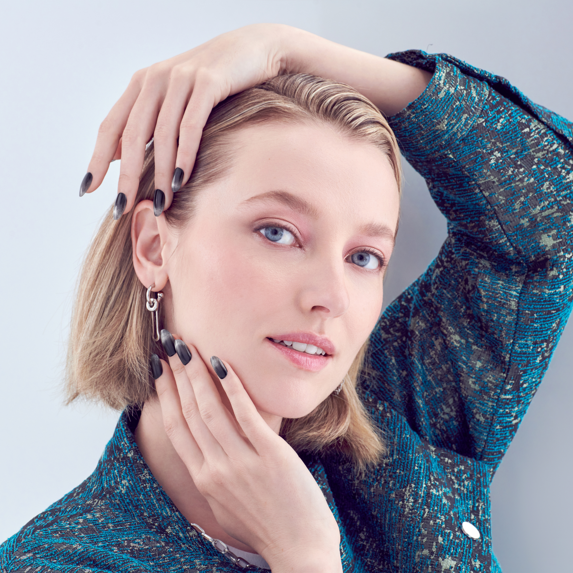 Woman with short blonde hair wearing a blue and black textured jacket, showing black ombré press-on nails with a matte finish on both hands.