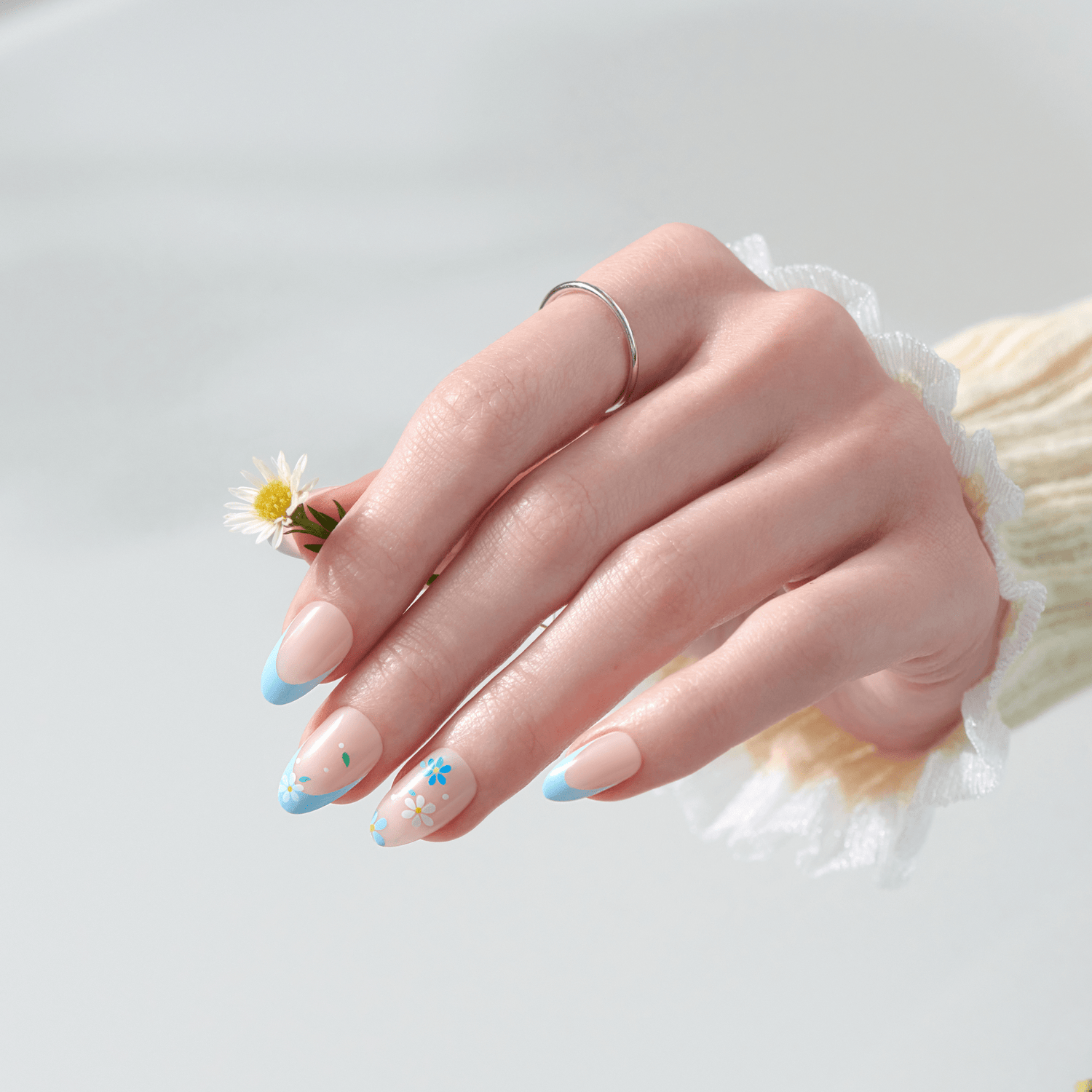 Manicured nails with blue tips and floral designs, holding small daisy flower.