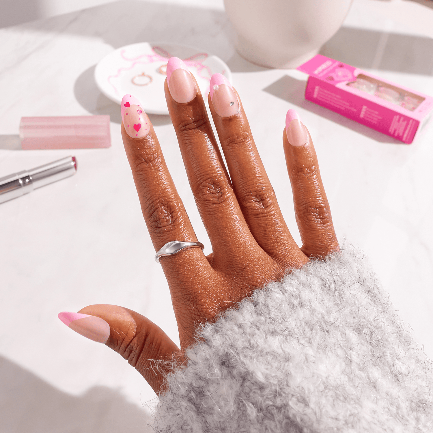 Hand displaying pink and white ombre nail polish with heart designs and rhinestones.