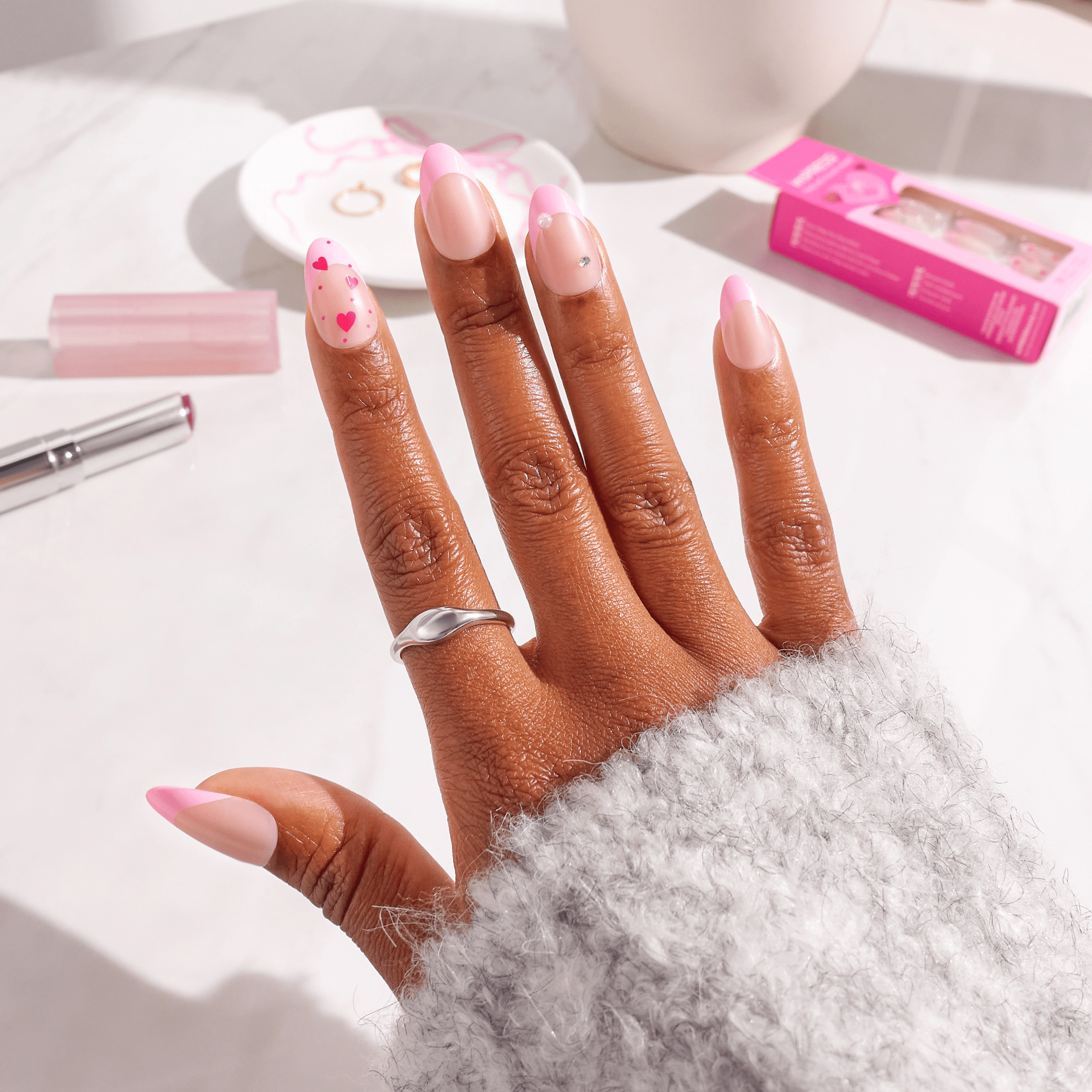Hand displaying pink and white ombre nail polish with heart designs and rhinestones.
