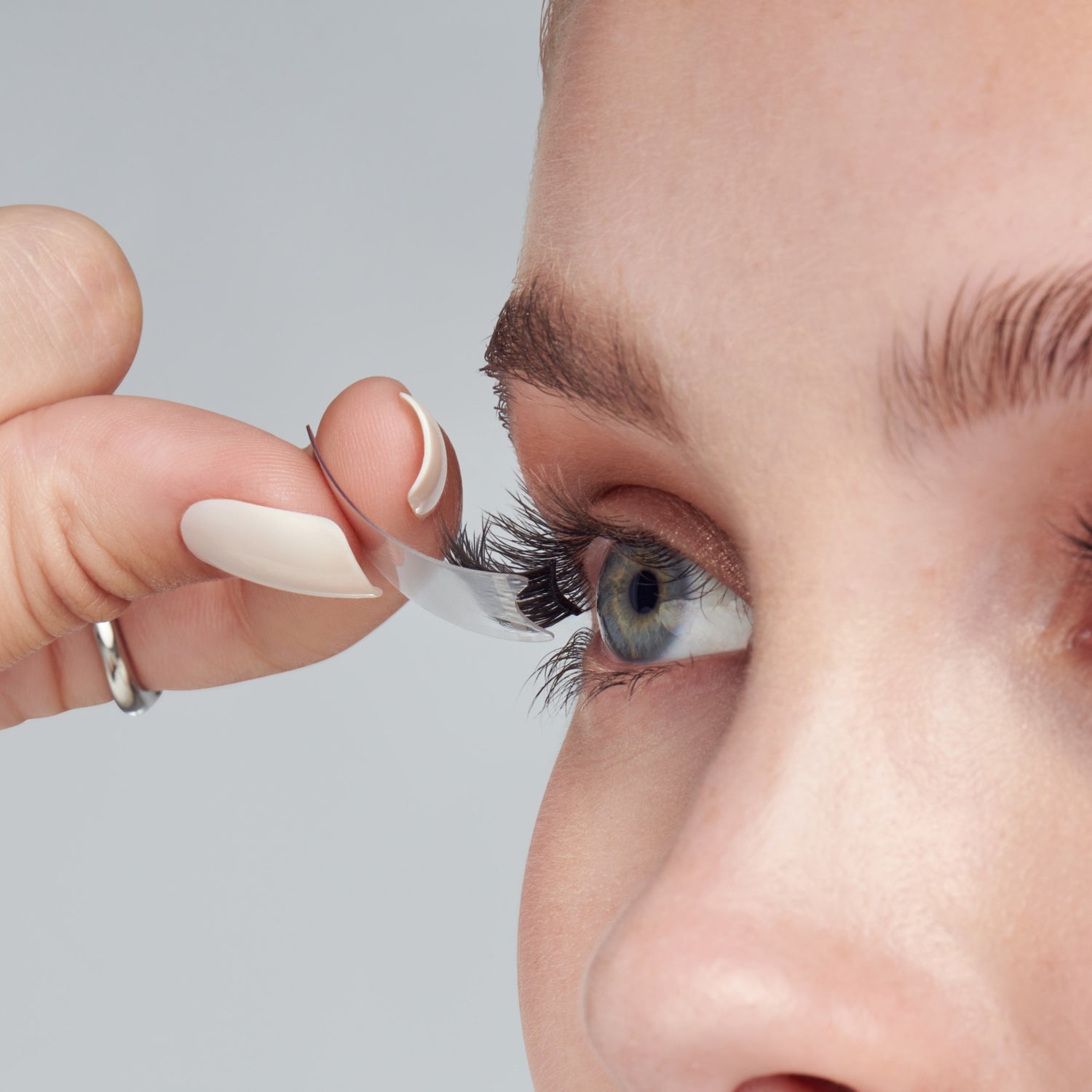 Close-up of a person’s eye with light skin and blue-green iris applying a clear self-adhesive false eyelash strip to the upper lash line.