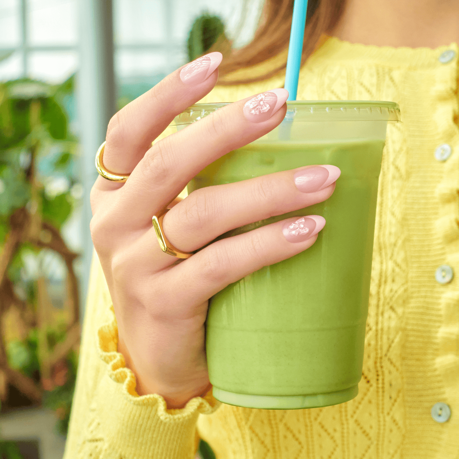 Woman holding green smoothie drink wearing yellow sweater and gold rings