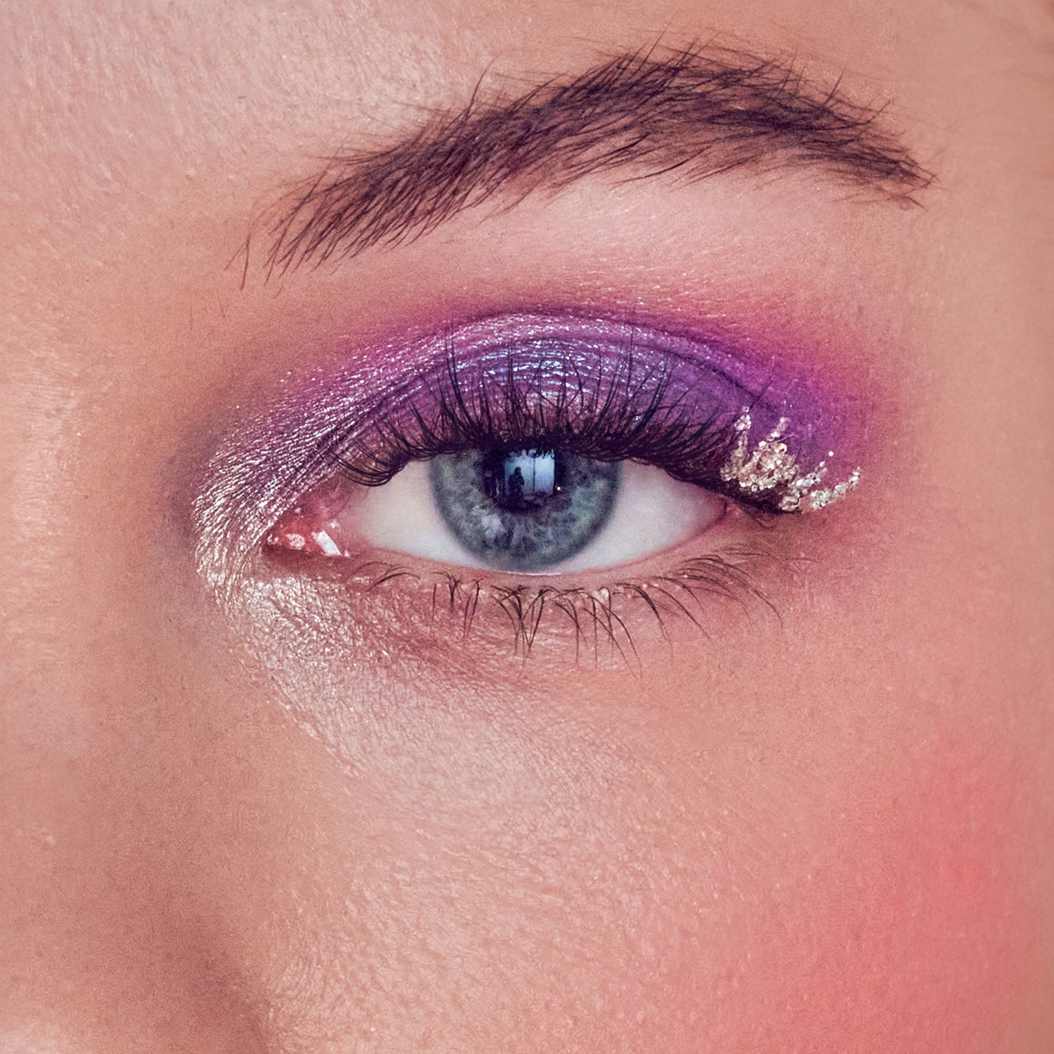 Close-up of a blue eye with long, thick false eyelashes and shimmery purple eyeshadow accented by silver glitter on the outer corner.