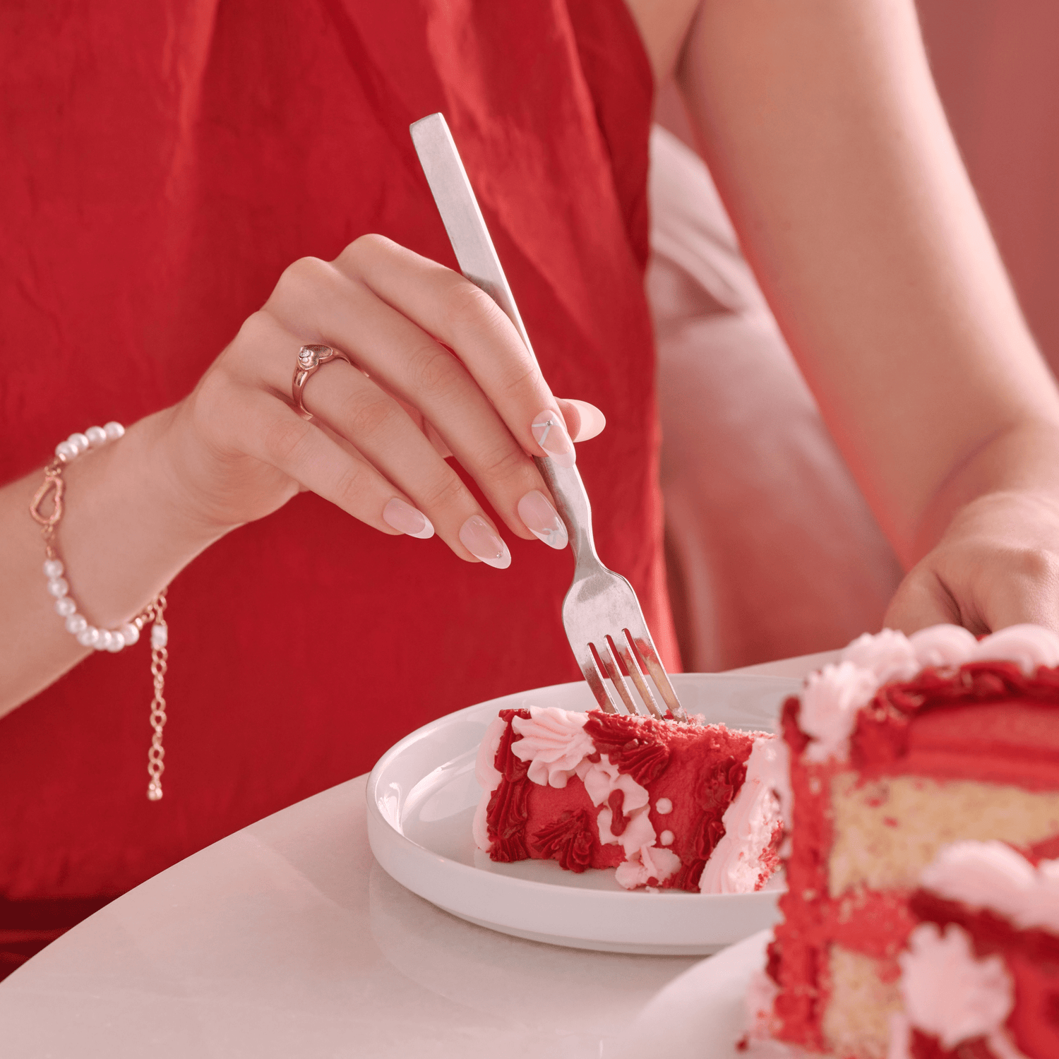 Hand with pale pink, heart-themed press-on nails holding a fork cutting a slice of red velvet cake on a white plate, person wearing red dress and pearl bracelet.