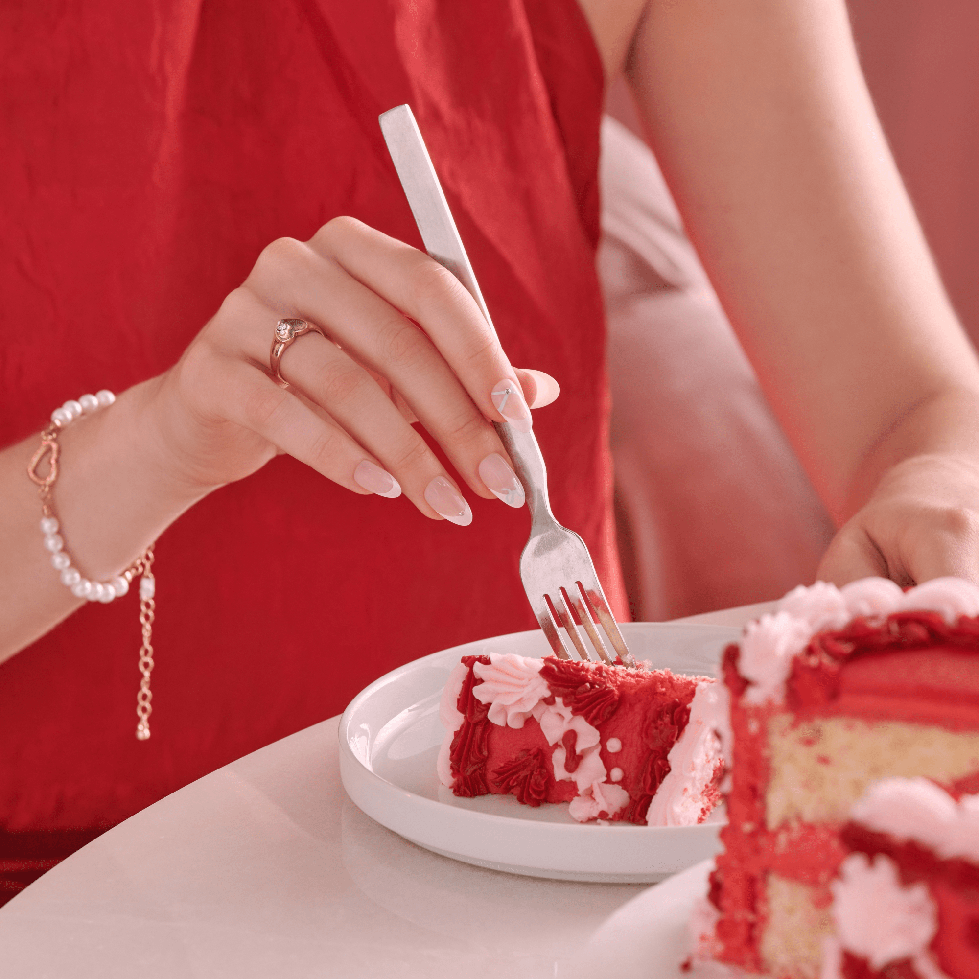 Hand with pale pink, heart-themed press-on nails holding a fork cutting a slice of red velvet cake on a white plate, person wearing red dress and pearl bracelet.