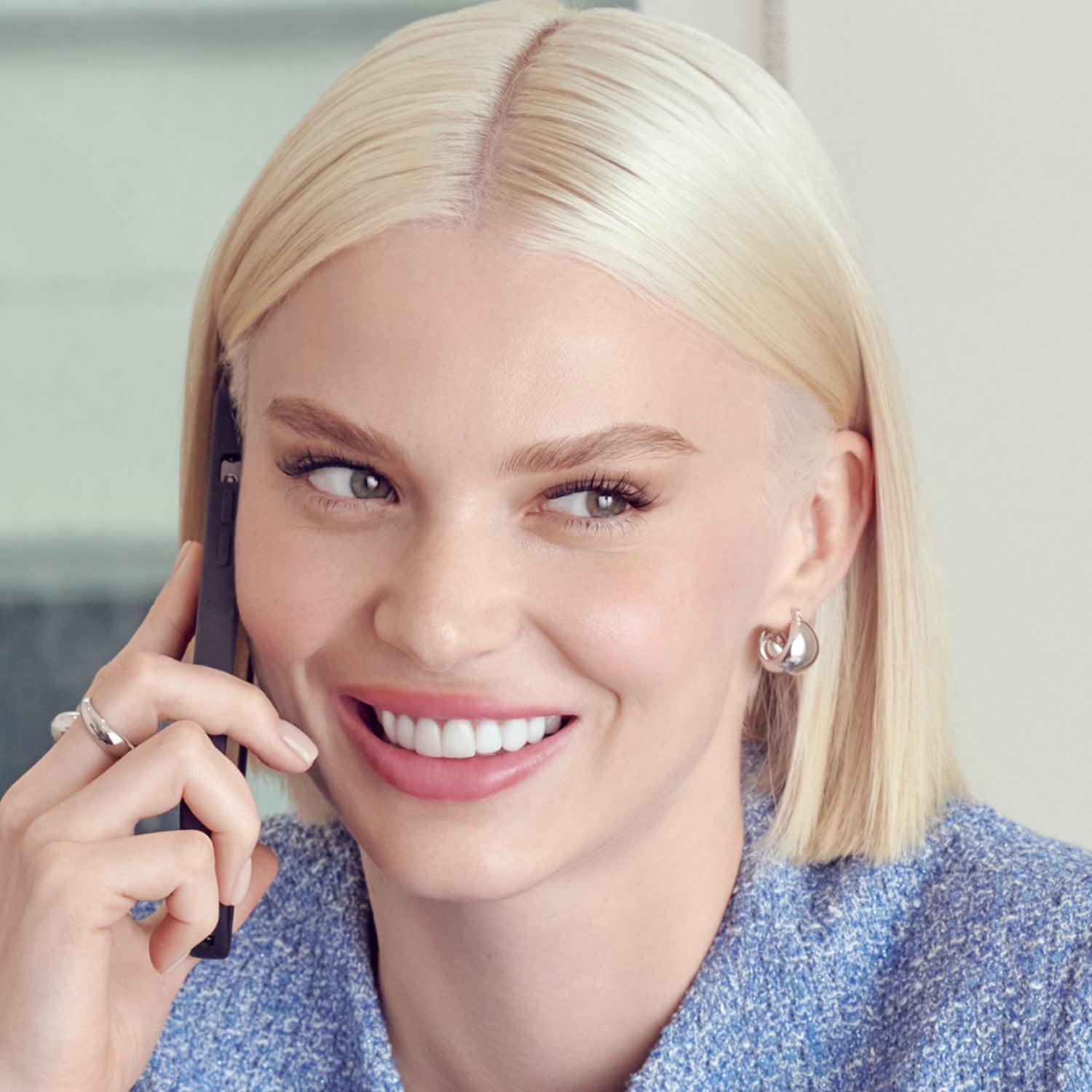 Close-up of a smiling woman with straight blonde hair wearing silver earrings and a blue textured top, showing natural-looking long eyelashes.