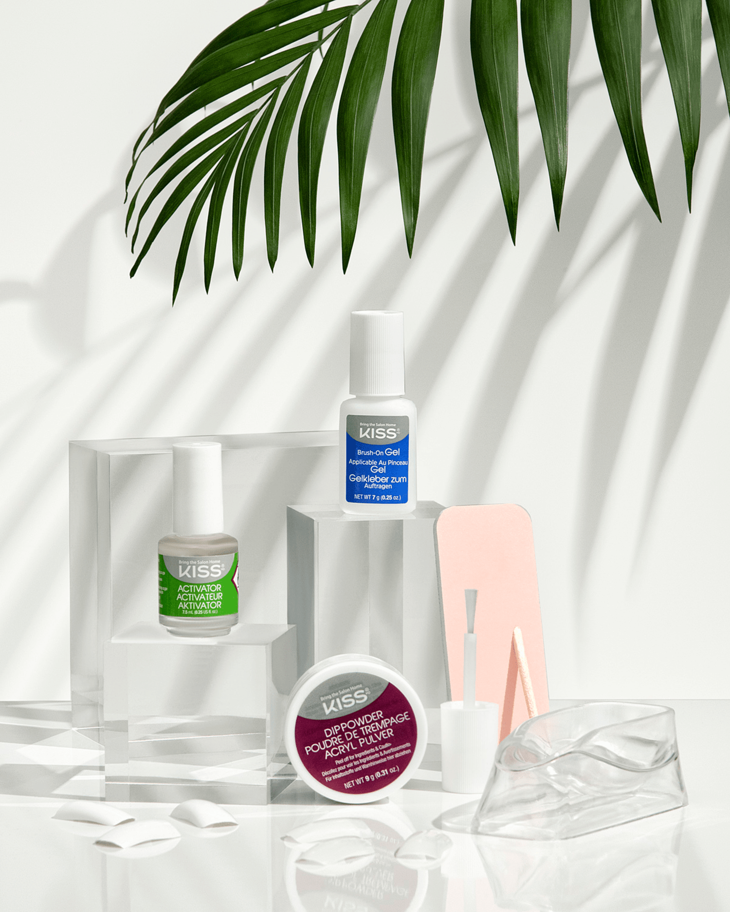 KISS Salon Dip Professional Dipping System showing activator bottle with green label, brush-on gel bottle with blue label, maroon dip powder jar, nail files, nail tips, and a clear finger soak bowl under a palm leaf shadow.