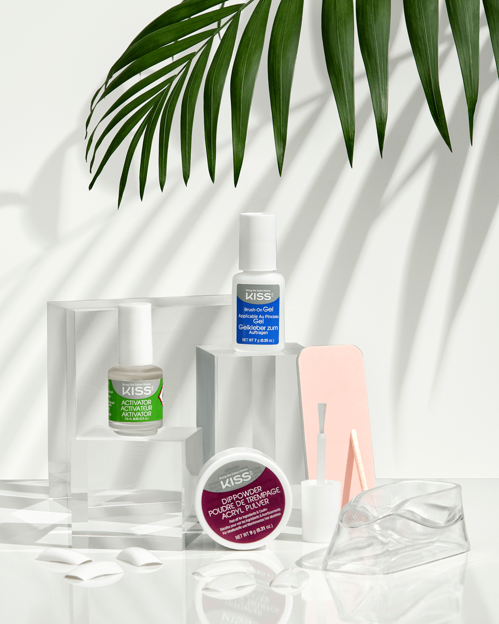 KISS Salon Dip Professional Dipping System showing activator bottle with green label, brush-on gel bottle with blue label, maroon dip powder jar, nail files, nail tips, and a clear finger soak bowl under a palm leaf shadow.
