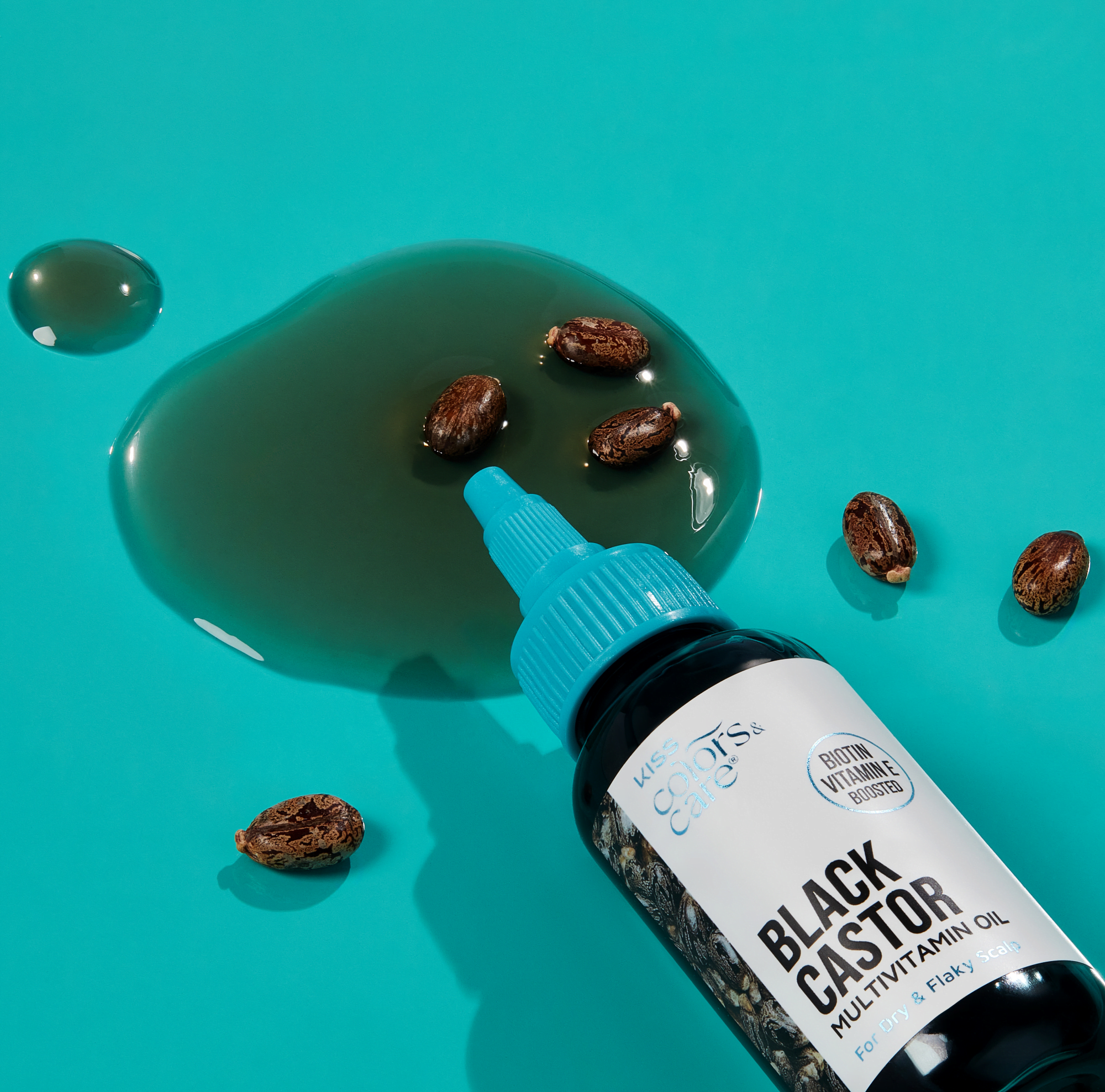 Bottle of KISS Colors &amp; Care Black Castor Multivitamin Oil with blue cap lying on teal surface with castor seeds and oil spill, labeled "Biotin Vitamin E boosted" for dry and flaky scalp.