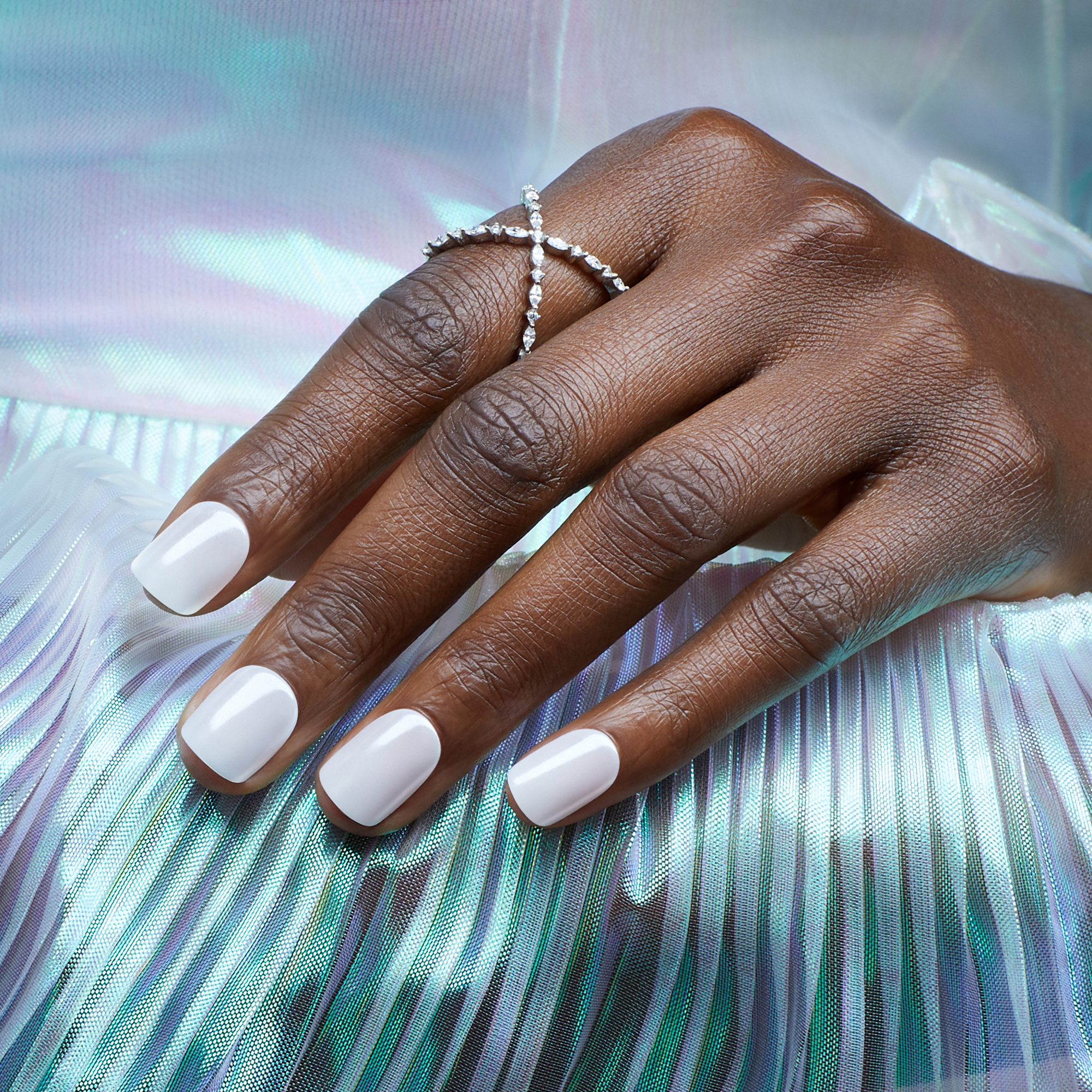 Dark-skinned hand with glossy, light gray press-on nails wearing a delicate silver crystal ring, resting on iridescent, pleated fabric.