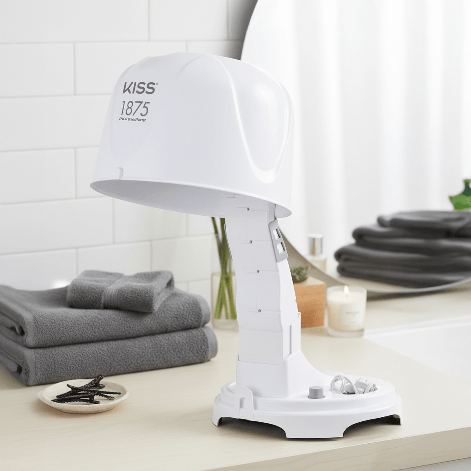 White KISS 1875W Salon Bonnet Hair Dryer on a bathroom counter with gray towels, hair clips, and a lit candle in the background.