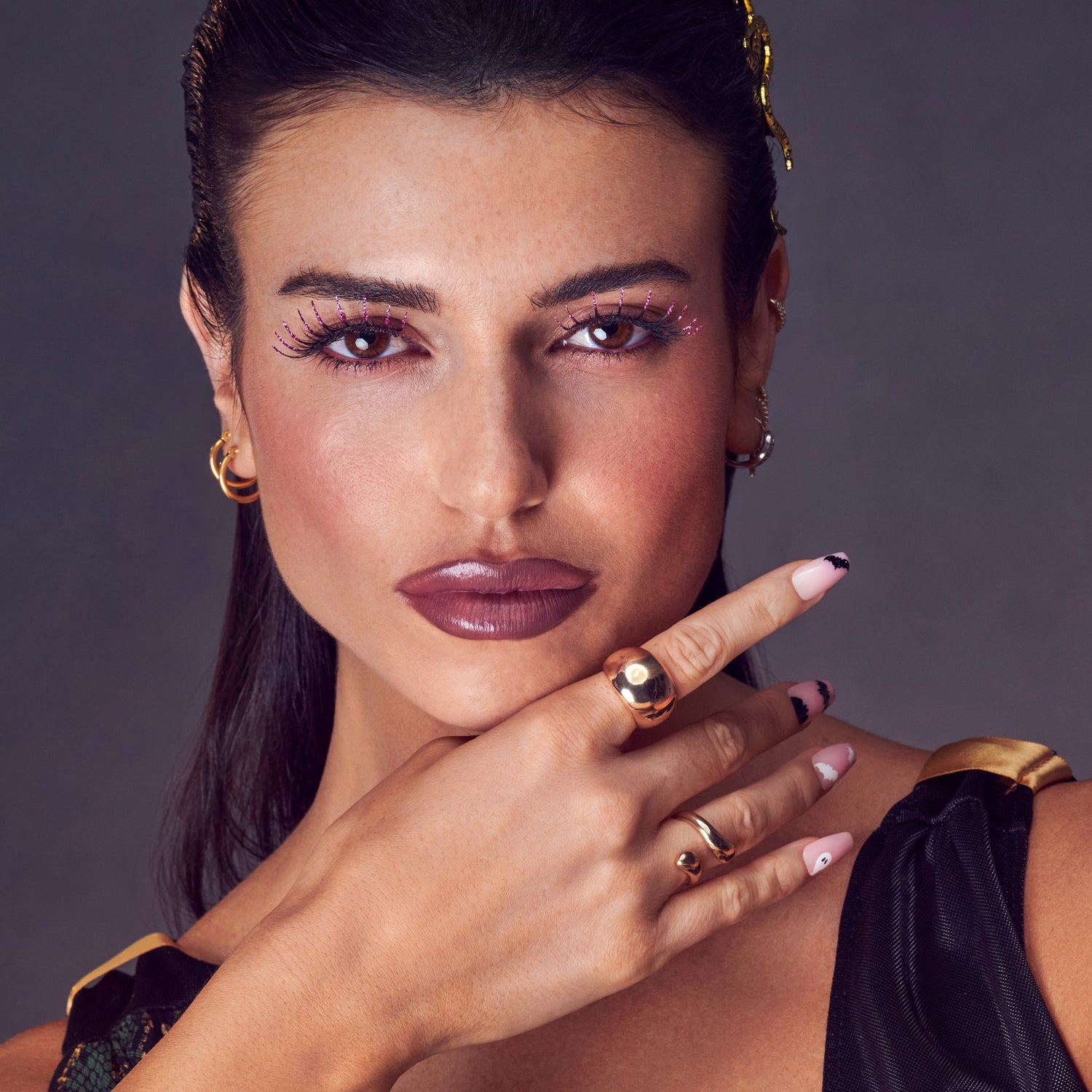 Close-up of a woman with dark hair wearing purple decorated false eyelashes with sparkle accents, mauve lipstick, and Halloween-themed nail art featuring bats and ghosts. She wears gold rings and a black outfit with gold trim against a neutral background.