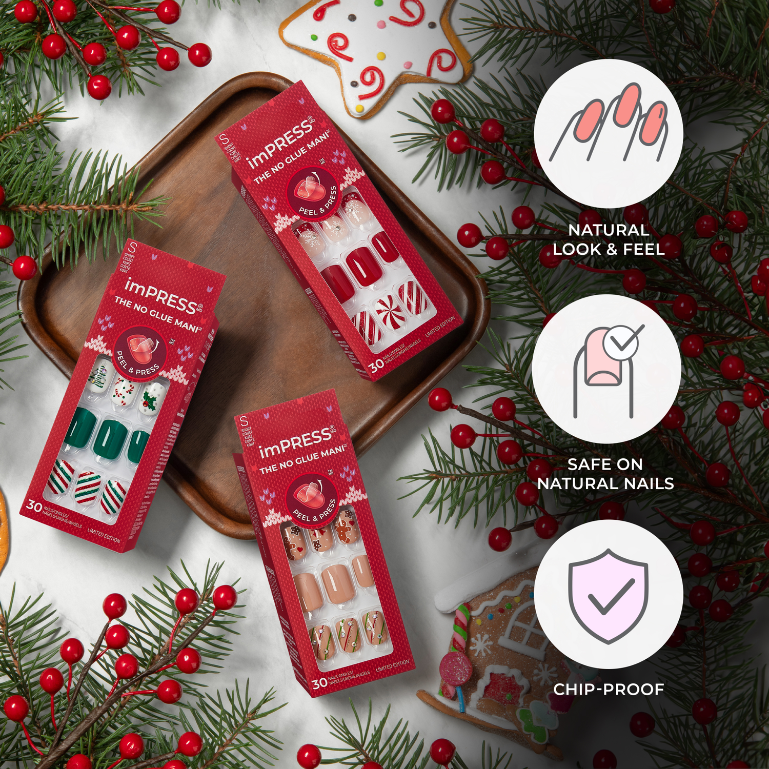 Three red-packaged imPRESS Press On Nails gift sets with holiday-themed nail designs on a wooden tray and marble surface, surrounded by festive greenery and cookies. Text highlights \