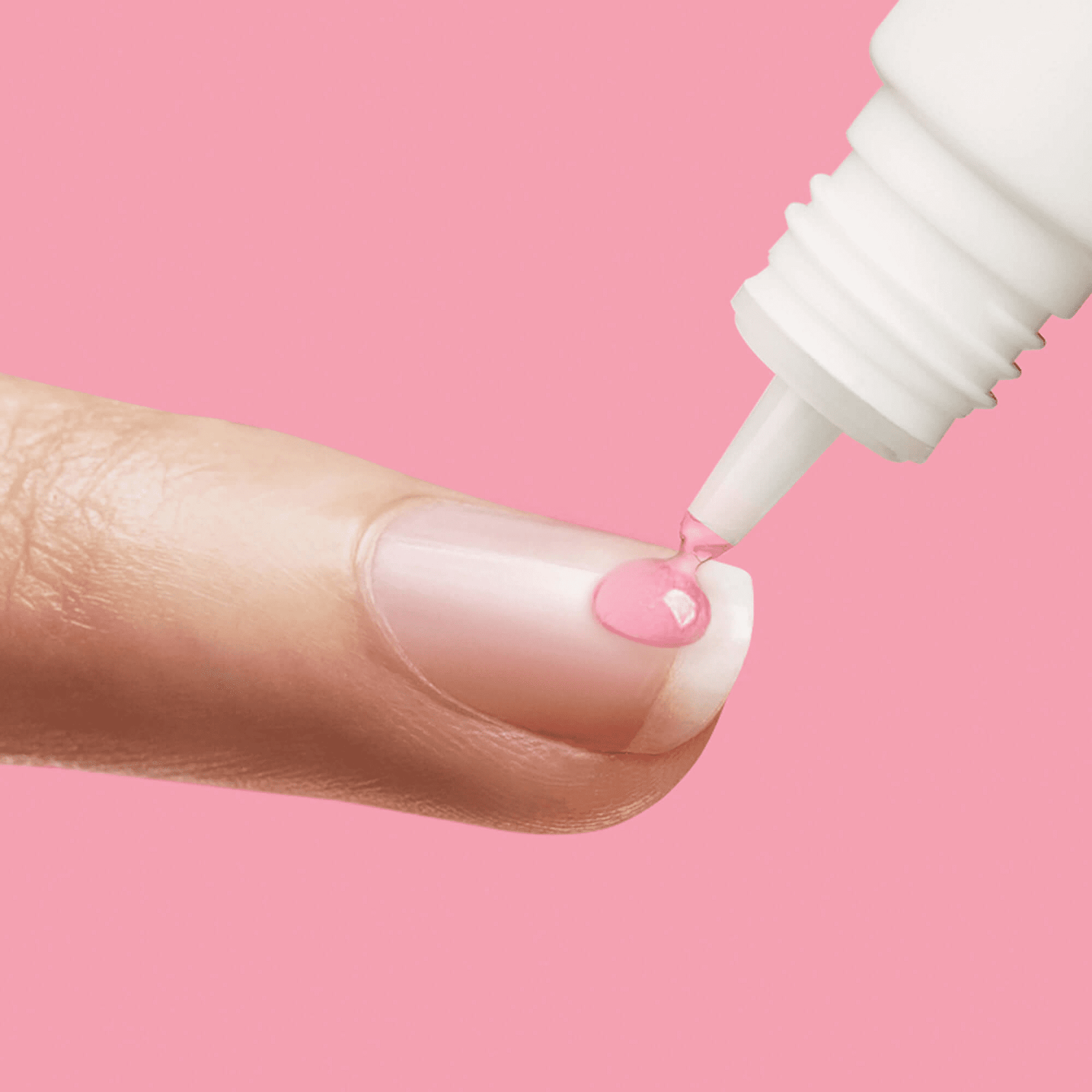 Close-up of a natural fingernail with a drop of pink nail glue being dispensed from a white tube applicator against a pink background.