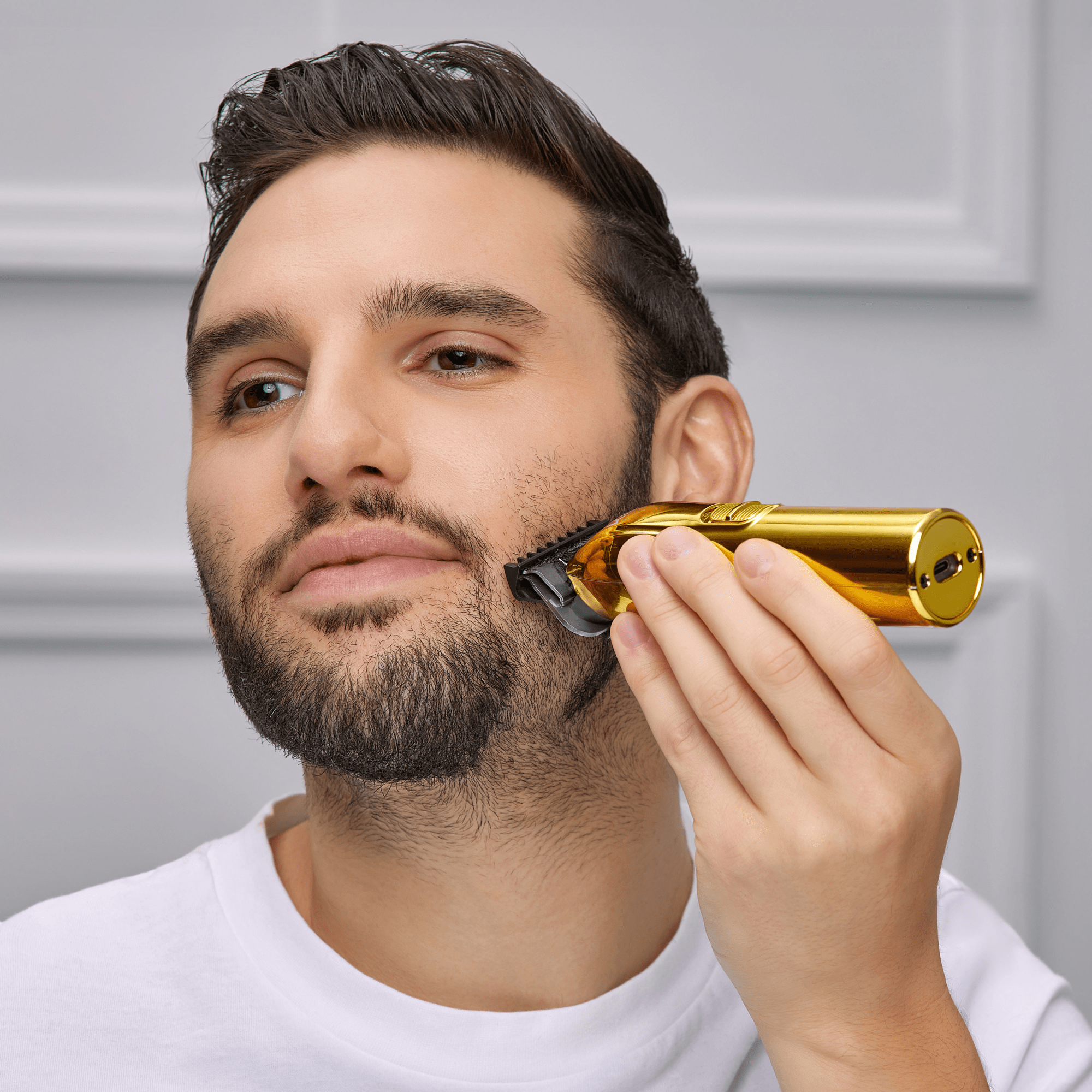 Man with short dark hair and beard uses a shiny gold cordless T-blade beard and hair trimmer to groom his cheek, wearing a white shirt against a gray background.