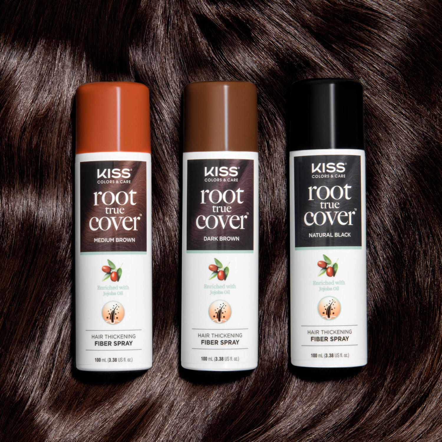 Three KISS Colors &amp; Care Root True Cover hair thickening fiber spray cans in Medium Brown, Dark Brown, and Natural Black, on a dark brown hair background, each labeled as enriched with jojoba oil.
