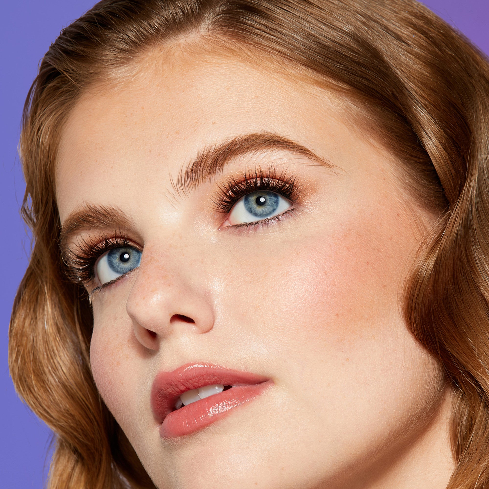 Close-up of a young woman with blue eyes and natural makeup showcasing long, full lashes against a solid purple background, highlighting the effect of false eyelashes.