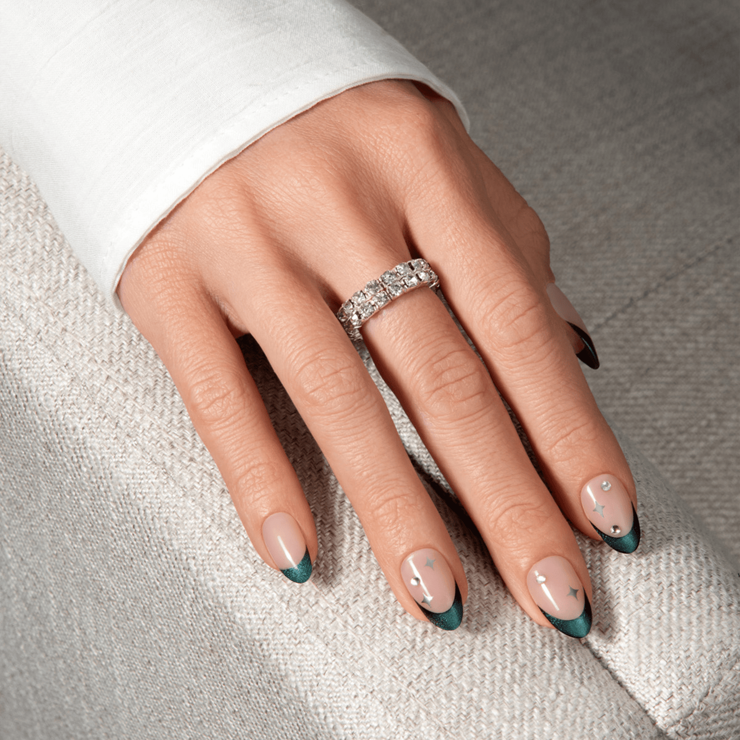 Hand with medium-length almond-shaped press-on nails featuring a nude base, dark teal French tips, and small silver star and gem accents, against a textured grey fabric background.