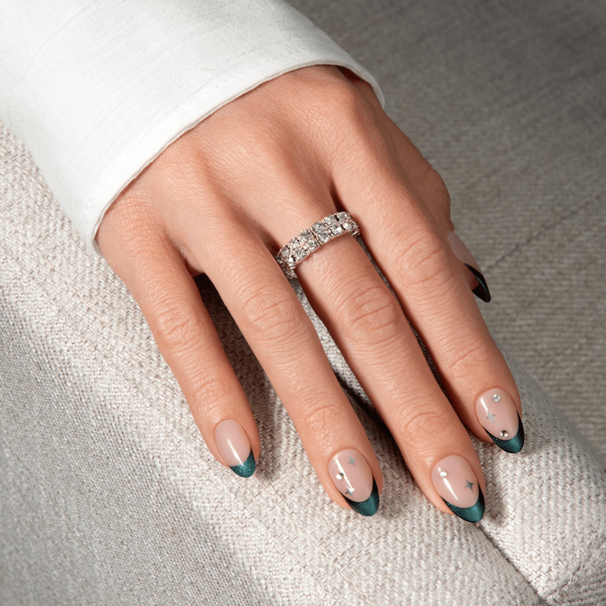 Hand with medium-length almond-shaped press-on nails featuring a nude base, dark teal French tips, and small silver star and gem accents, against a textured grey fabric background.