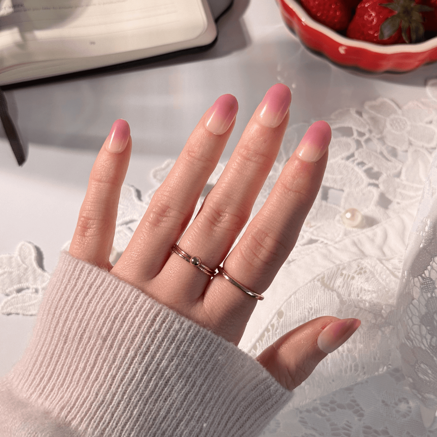 Hand with ombré pink press-on nails worn with two thin rose gold rings, resting on white lace fabric beside a bowl of strawberries.