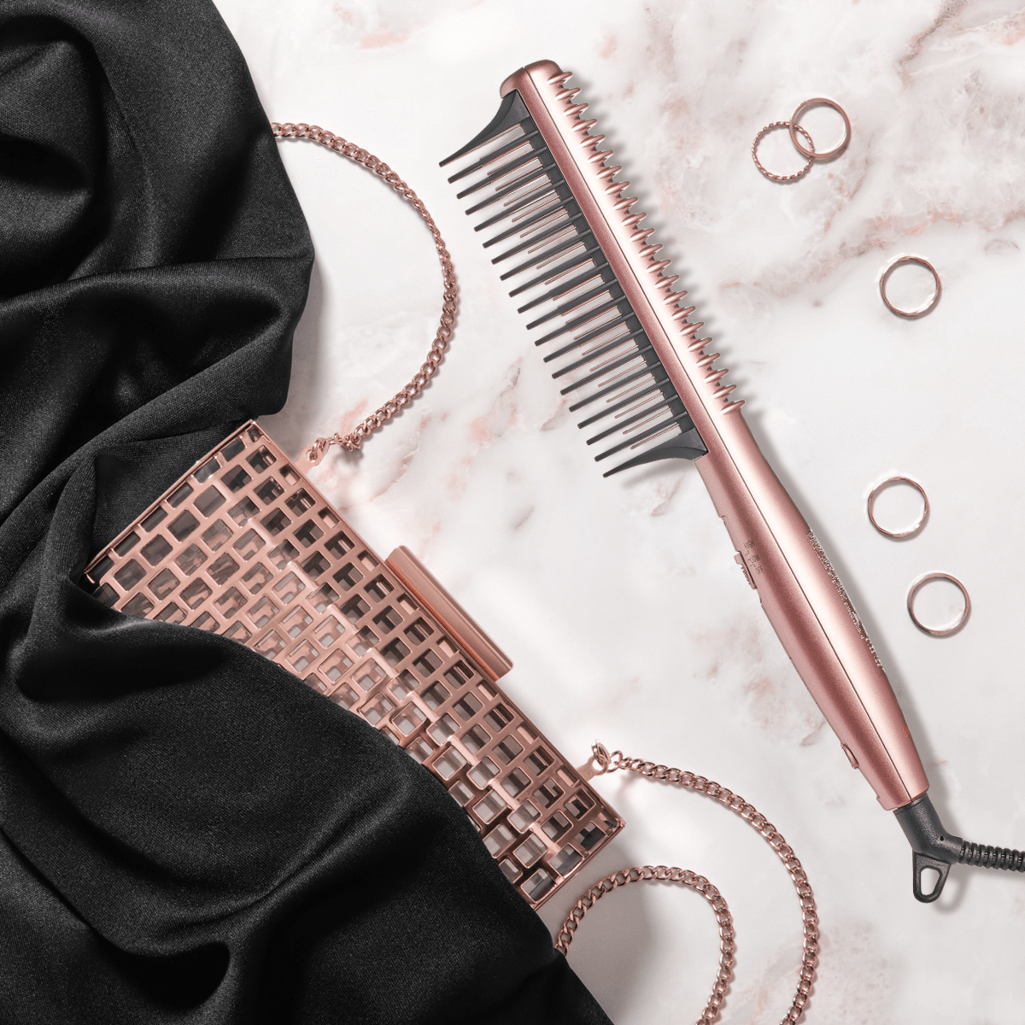 Sleek rose gold heat styling brush with black bristles lying on a marble surface next to a matching rose gold chain purse, black fabric, and four rose gold rings.