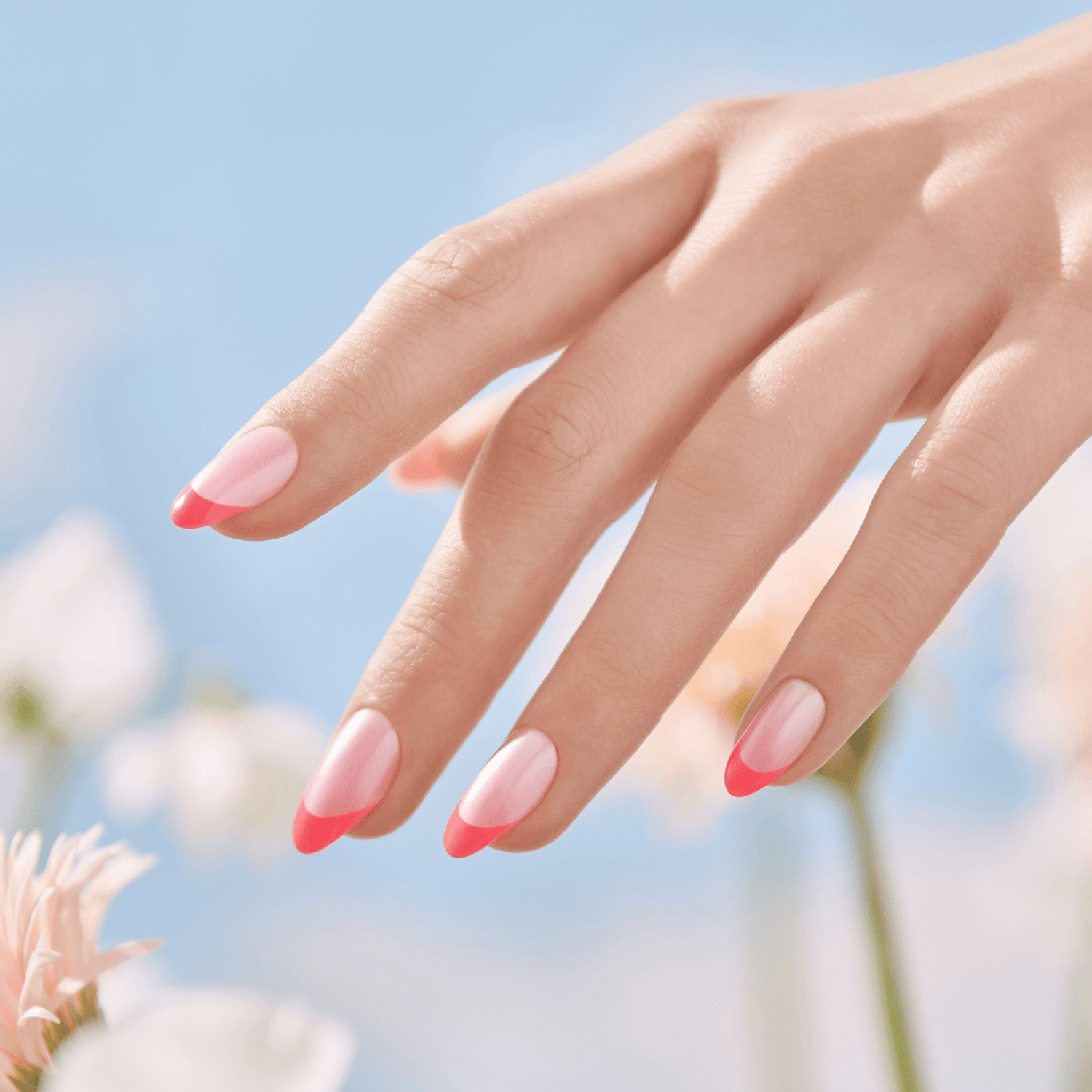 Hand with light pink press-on nails featuring bright coral-pink tips in a soft almond shape, set against a blurred floral background and clear blue sky.