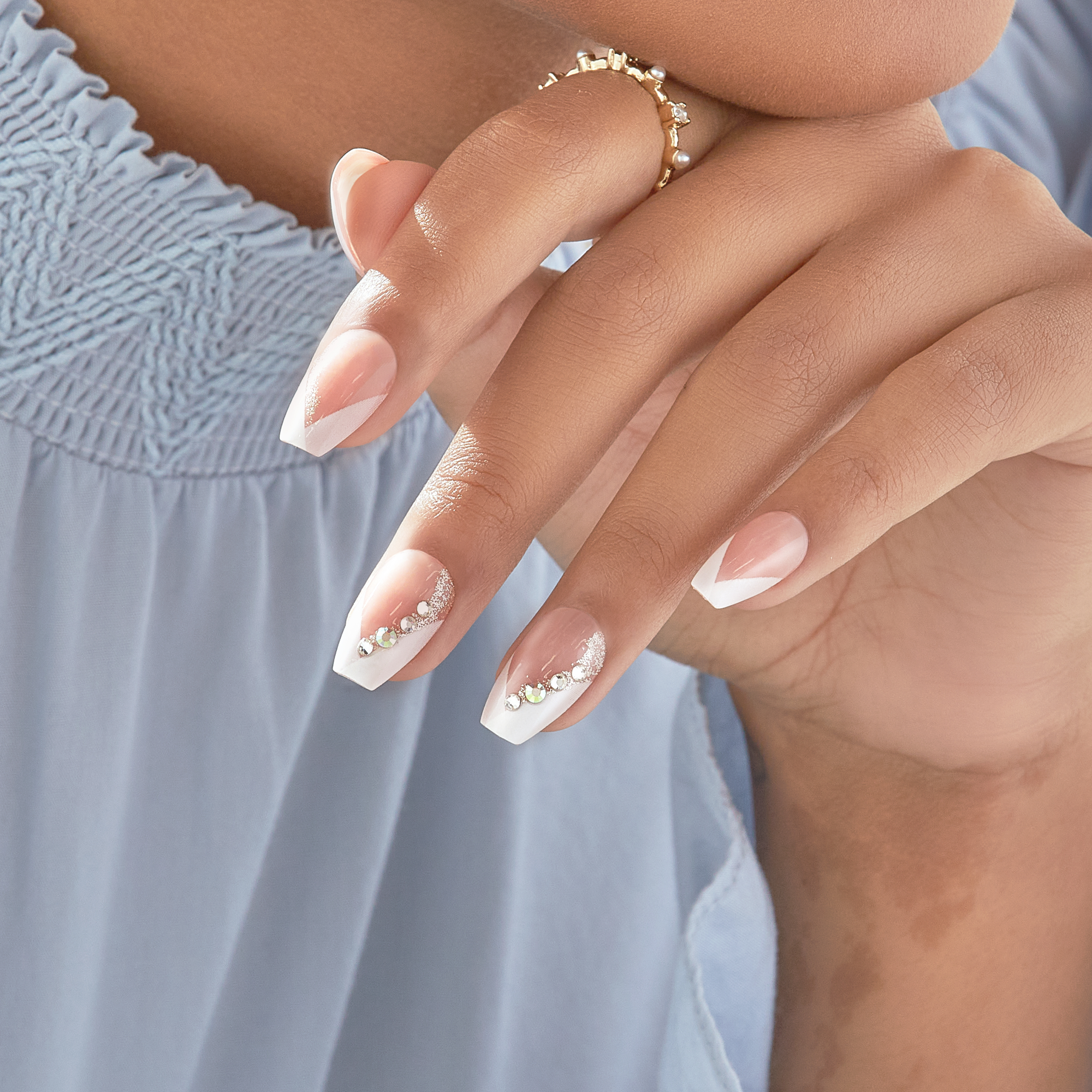 Hand with medium-length coffin-shaped press-on nails in a French manicure style with white tips and subtle glitter and rhinestone accents on two nails, against a light blue gathered fabric background.