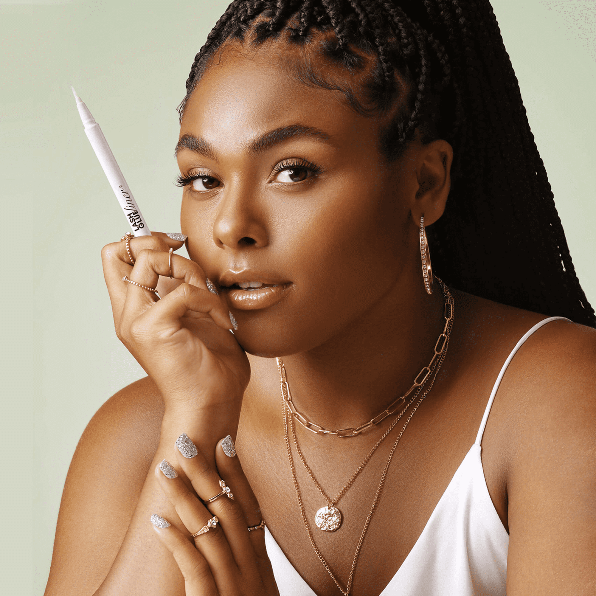 Woman with braided hair wearing layered gold necklaces, hoop earrings, and a white tank top holds a white pen-style KISS Lash GLUEliner with clear adhesive.