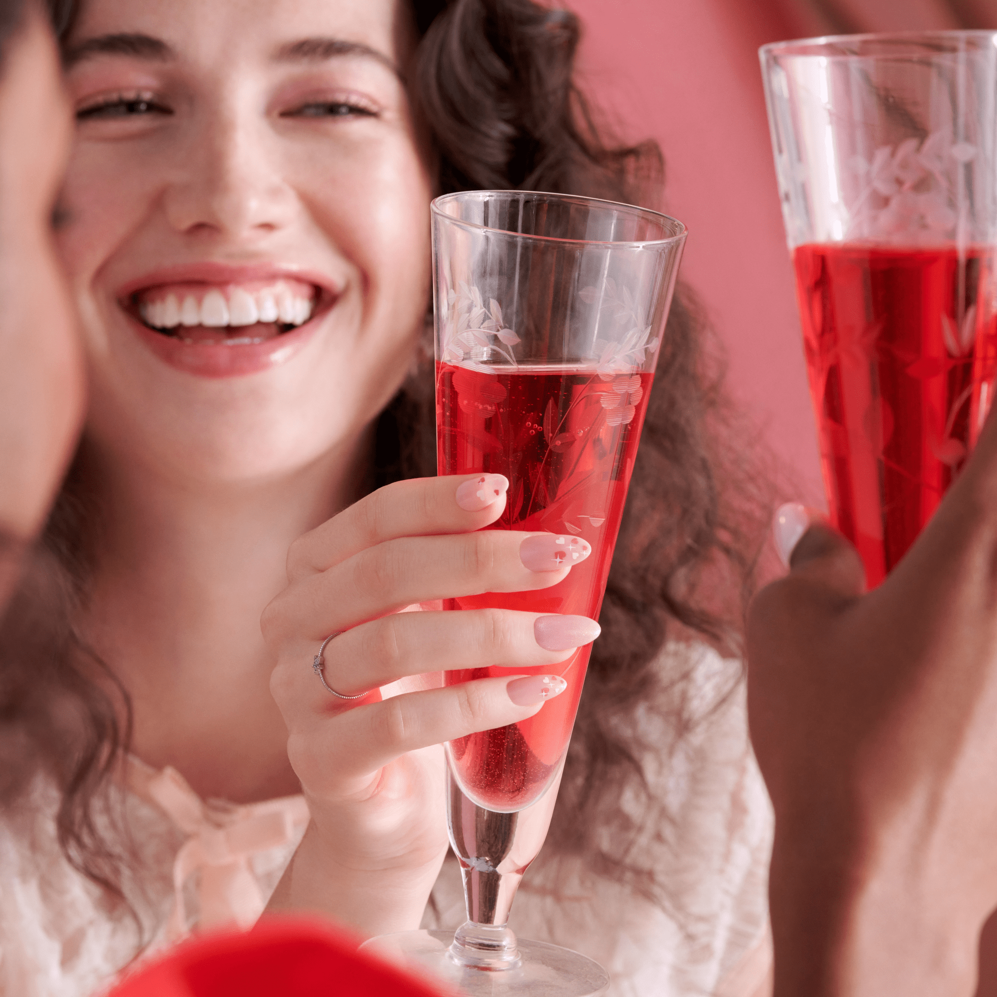 Smiling woman holding a decorated champagne flute with light pink press-on nails featuring small red heart and cross designs, toasting with another person.