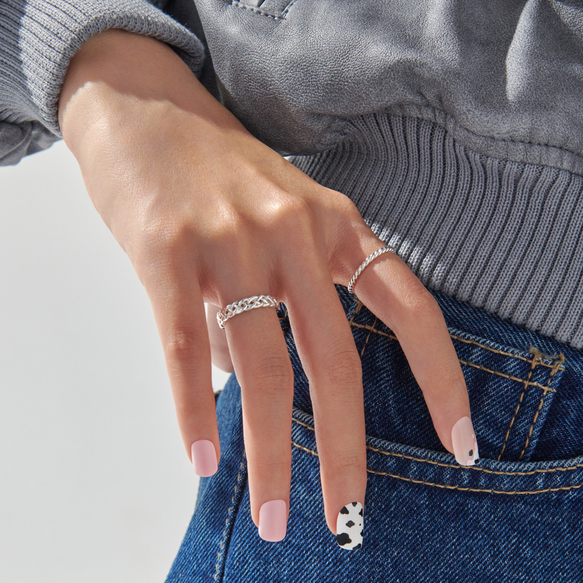 Hand with light pink press-on nails, two featuring black and white cow print accents. Wearing silver rings and resting on blue denim.