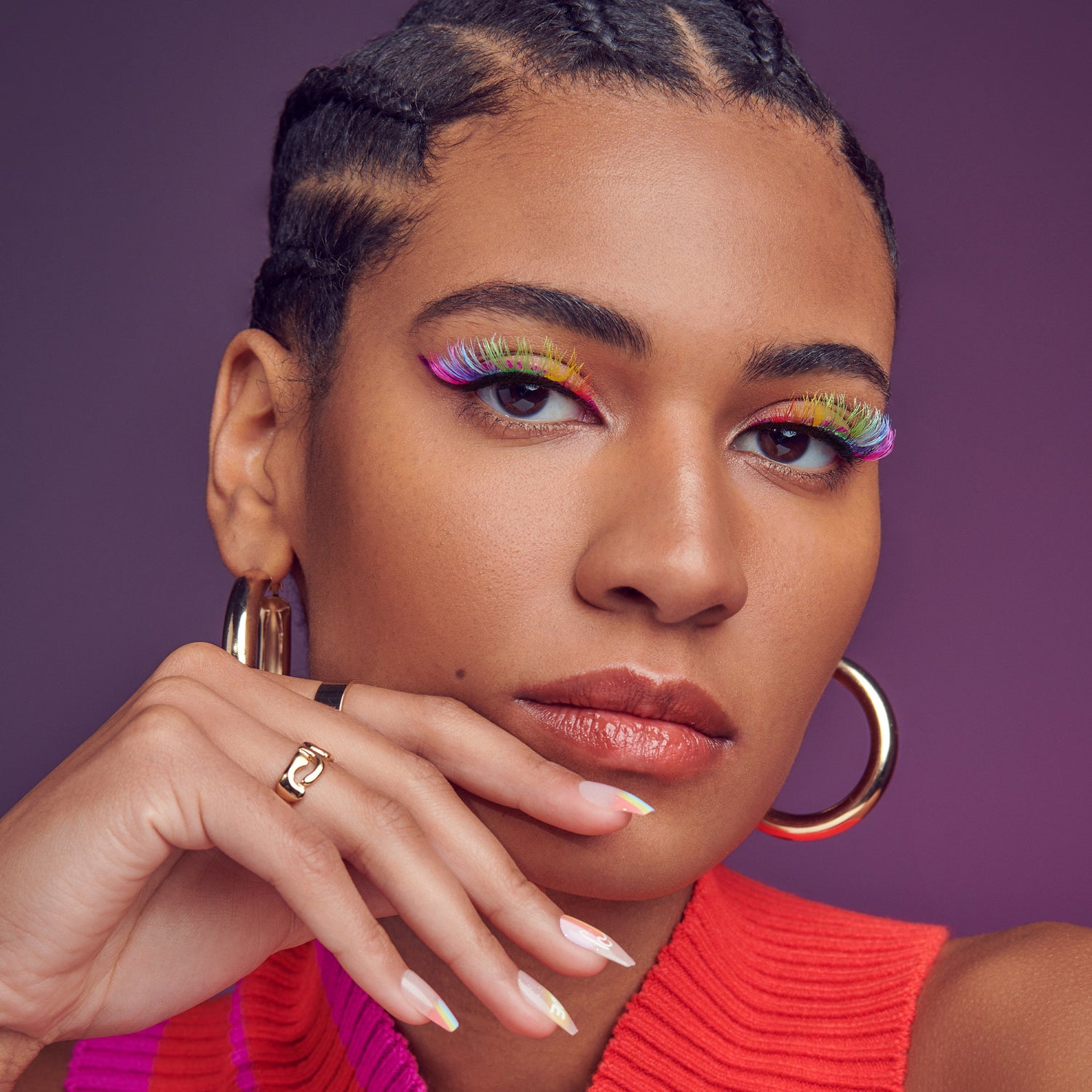Close-up of a woman wearing colorful rainbow false eyelashes that transition through pink, purple, blue, green, yellow, and red shades, with glossy lips, gold hoop earrings, and a pink and orange sleeveless top against a purple background.