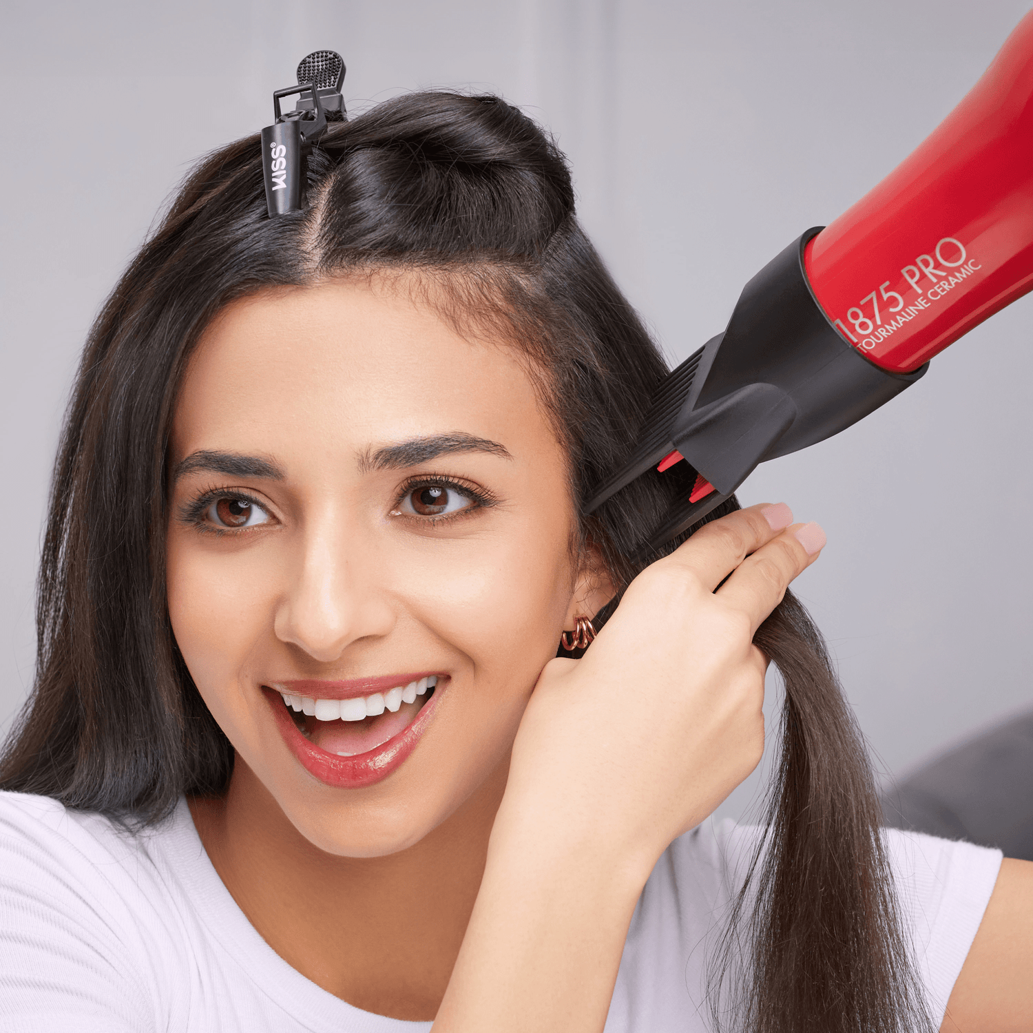 Smiling woman using a red and black 1875 Pro Tourmaline Ceramic Dryer on her straight dark hair, secured with a black KISS hair clip.