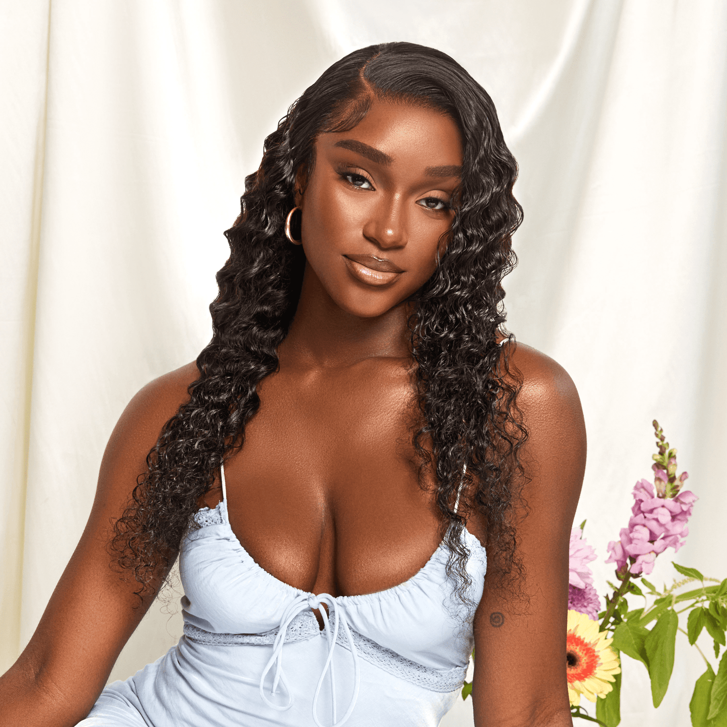 Model with long, curly black hair wearing a light blue spaghetti strap top, posing against a cream backdrop with pink and yellow flowers.