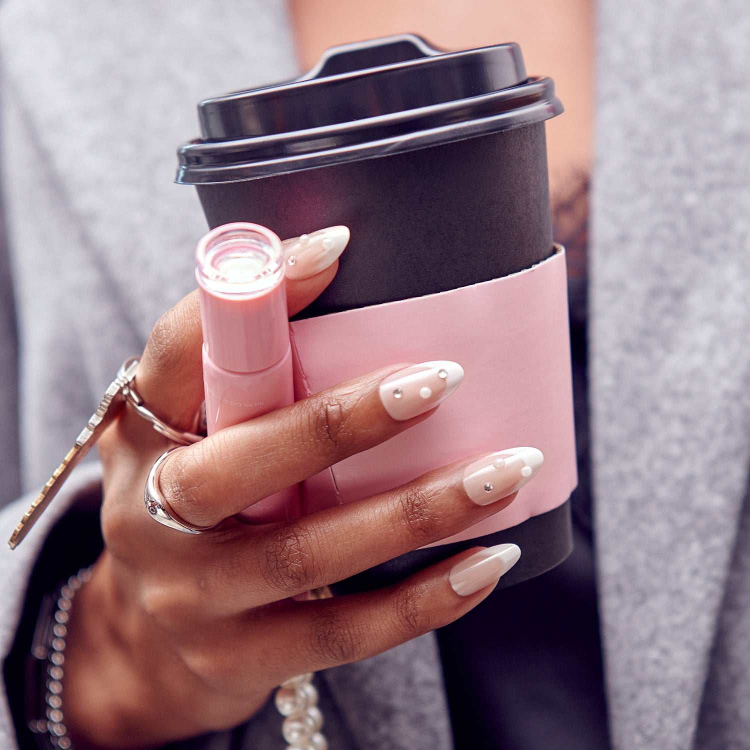 Person holding pink and black coffee cup with lip gloss, manicured nails with rhinestones.