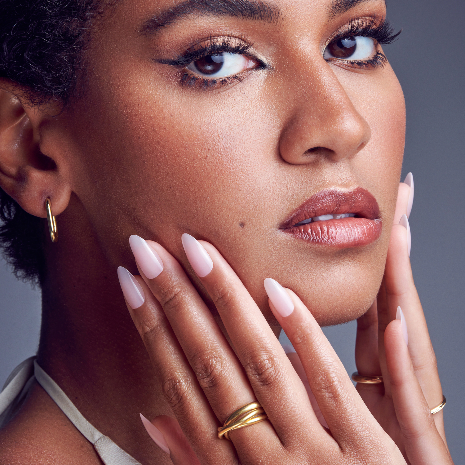 Close-up of a woman with medium skin tone showing long, almond-shaped press-on gel nails in a soft pink shade, touching her face gently, wearing gold rings and hoop earrings.
