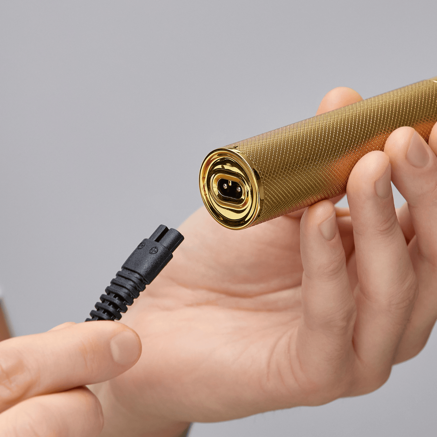 Hand holding a gold textured cylindrical cordless trimmer with a close-up on its charging port, while another hand holds a black charging cable near the connector.