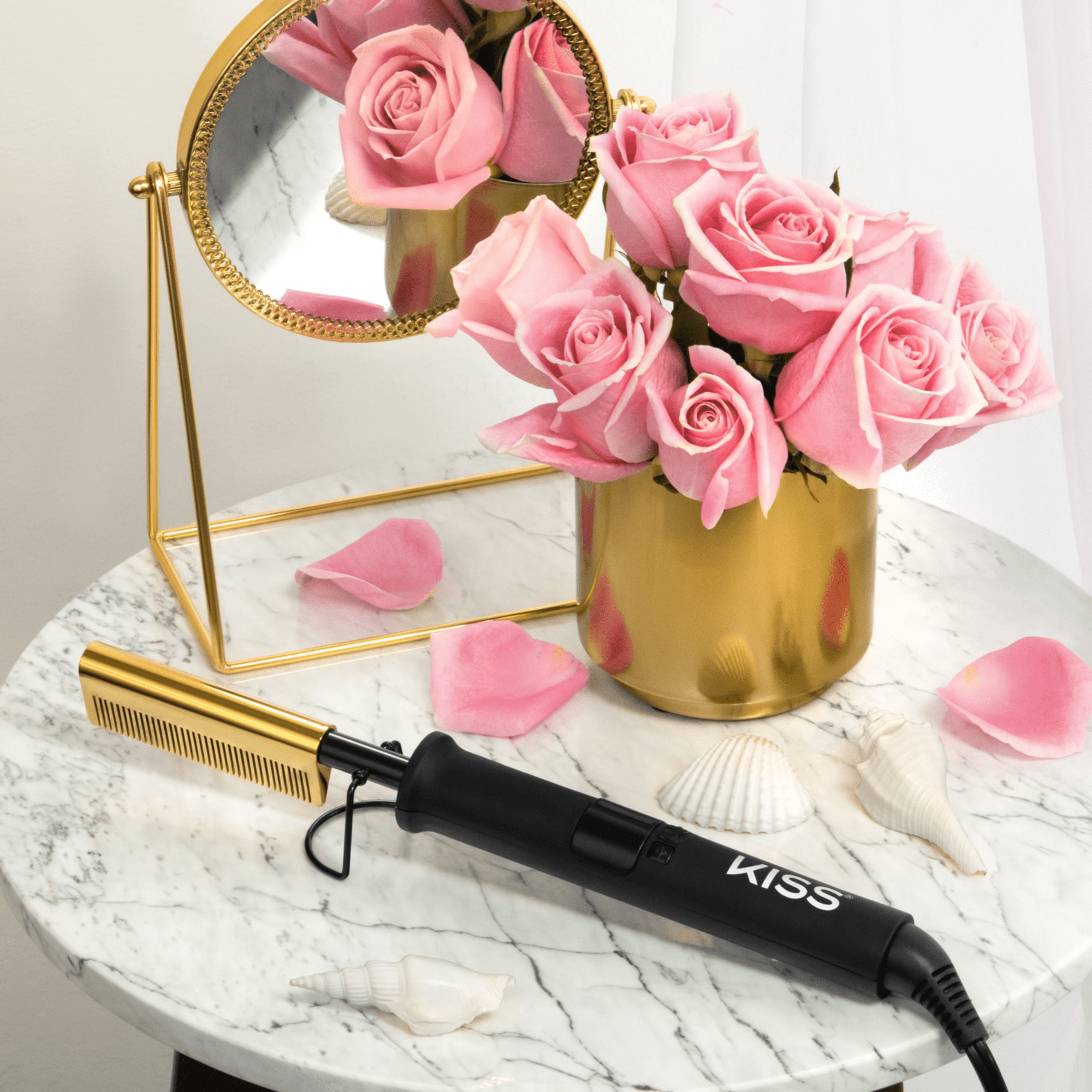 Black and gold KISS Professional Pressing Comb resting on a round white marble table with pink roses in a gold vase, seashells, and a small gold framed mirror.