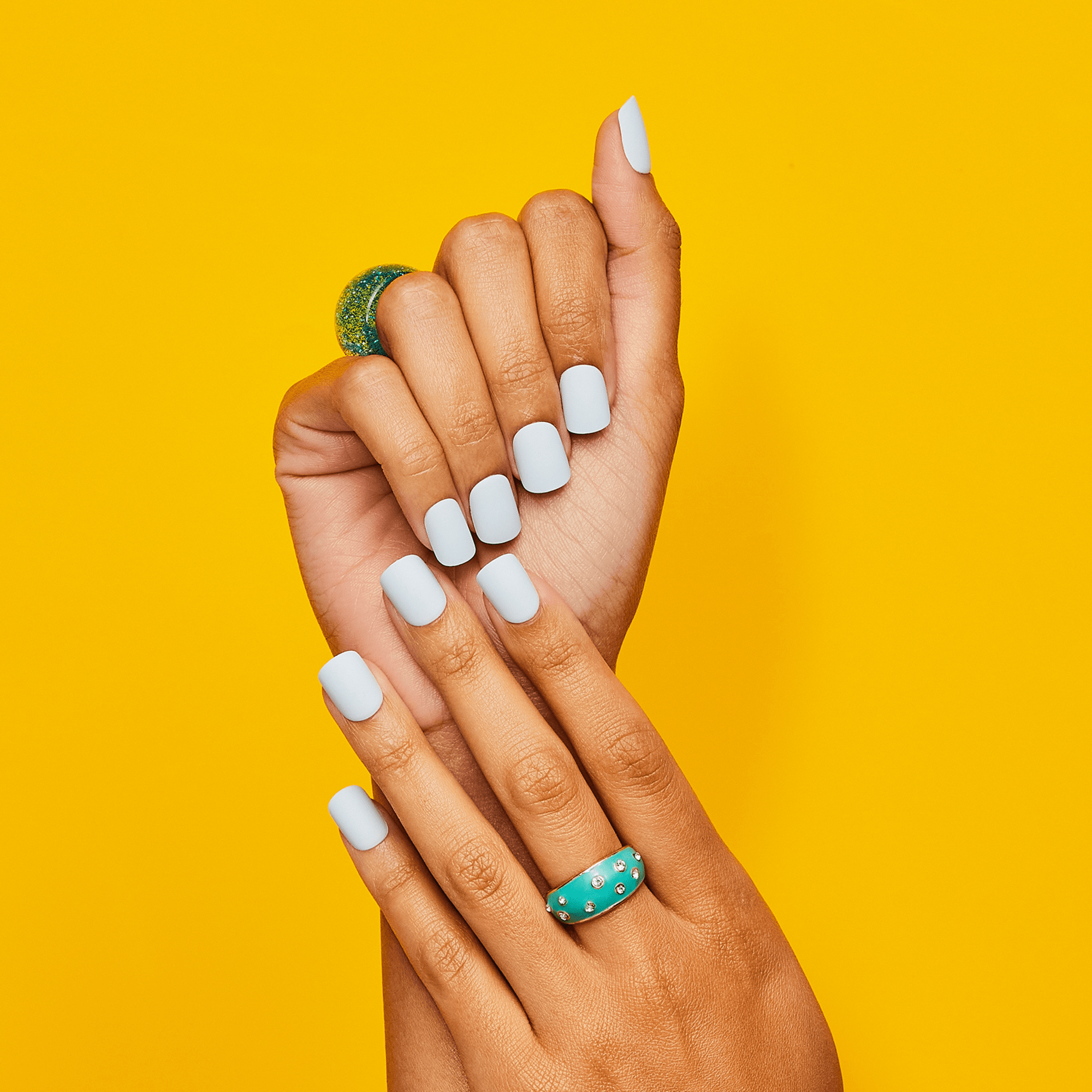 Two hands with medium brown skin tone display press-on nails in a soft matte pale blue color against a bright yellow background. The left hand features a glittery green ring and the right hand wears a turquoise ring studded with small gems.