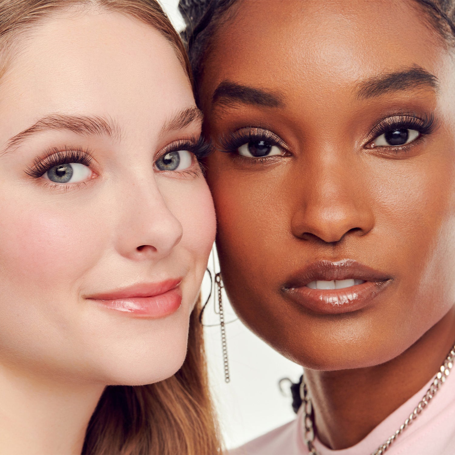 Two women with different skin tones posing closely together, both wearing makeup and jewelry.
