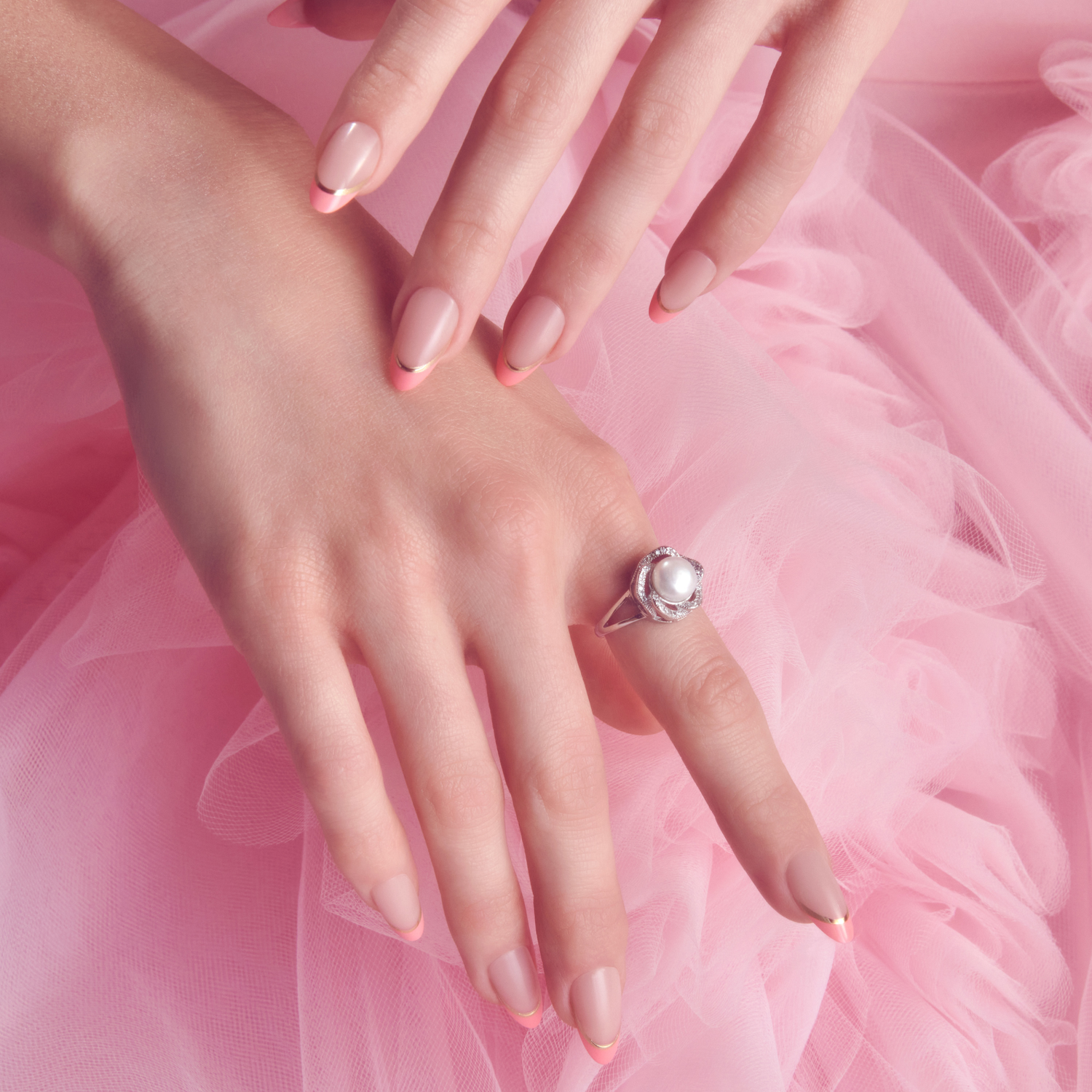 Hands with manicured nails featuring pink tips and thin gold accents, resting on pink tulle fabric; one hand wears a silver ring with a pearl center.