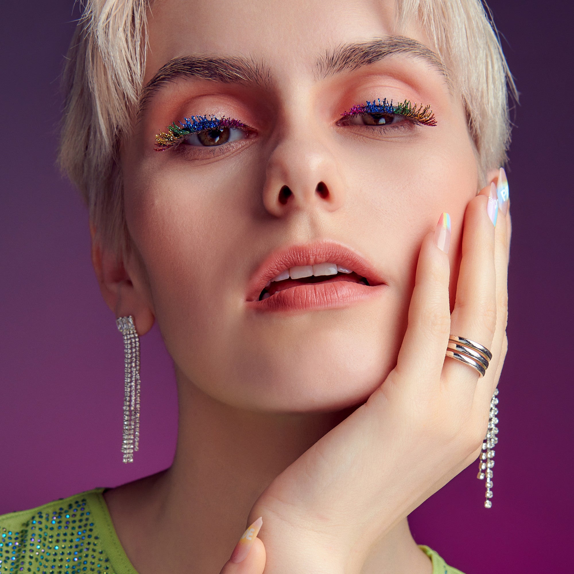 Close-up of a person with short blonde hair wearing rainbow-colored false eyelashes, sparkling dangling earrings, and rainbow-themed nail art on a purple background.
