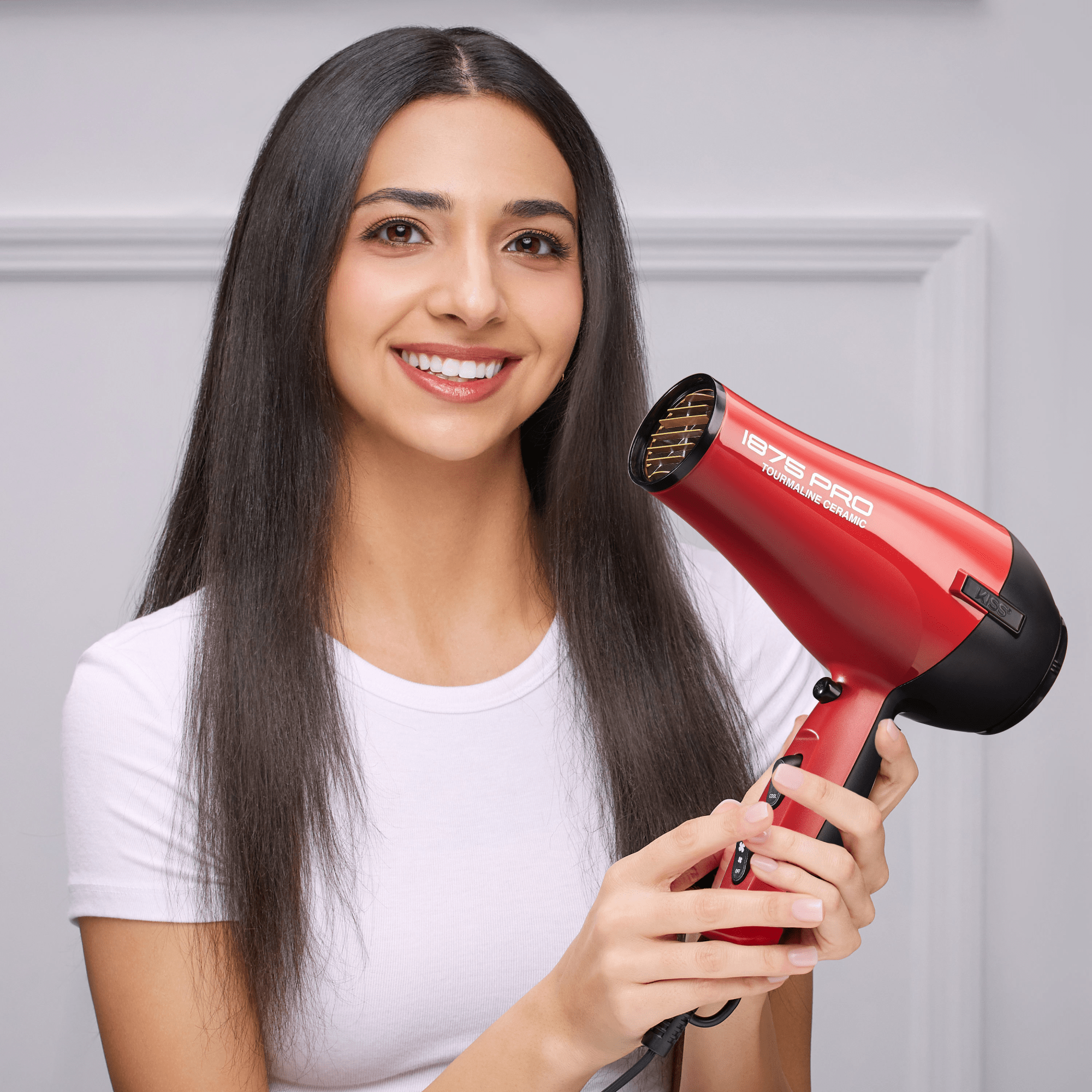 Smiling woman with long straight dark hair holds a red 1875 Pro Tourmaline Ceramic hair dryer, showing its controls and branding.